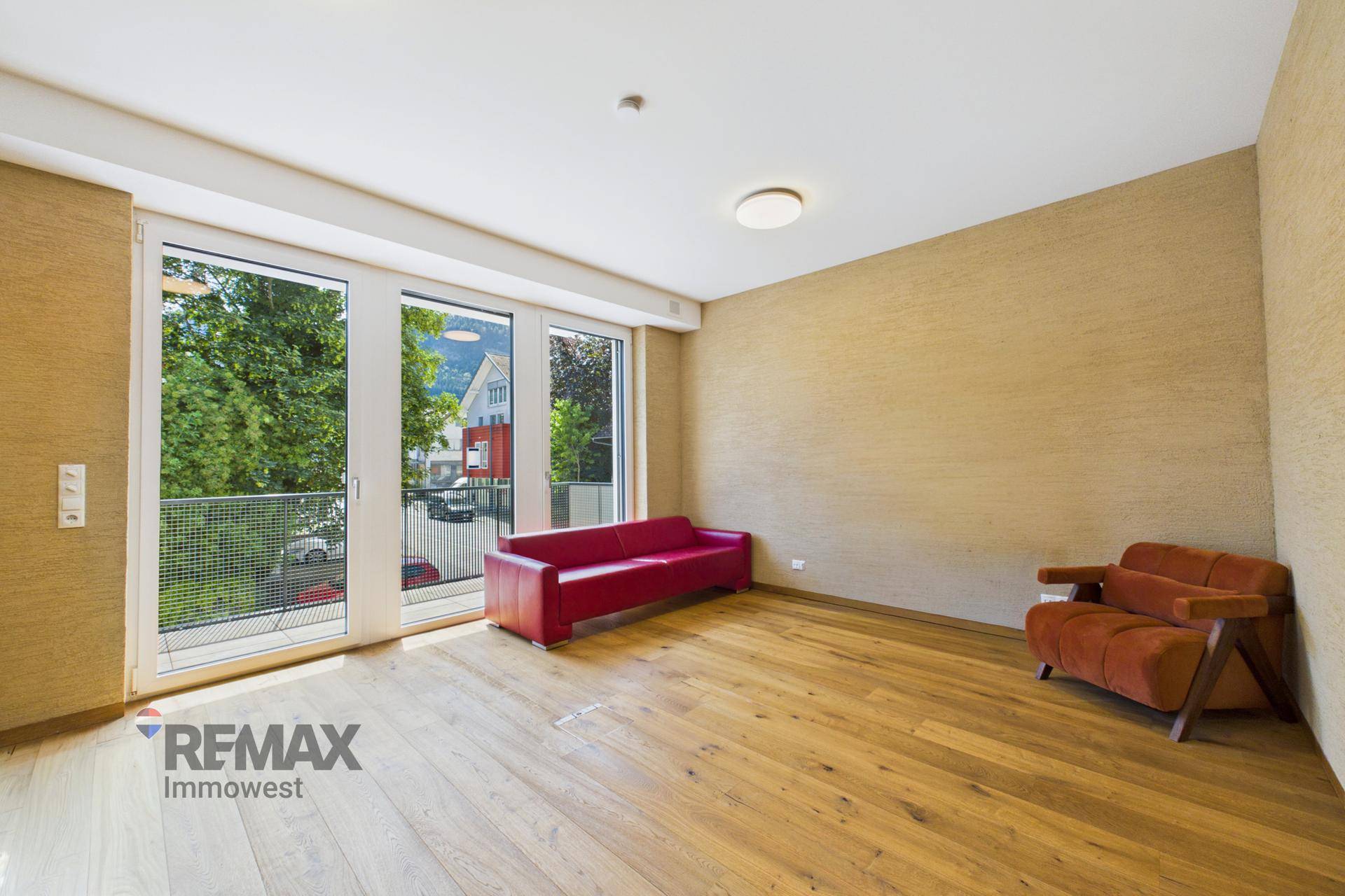 Heller Raum mit Holzplankenboden, großen Fenstern, einem roten Sofa und einem Sessel.