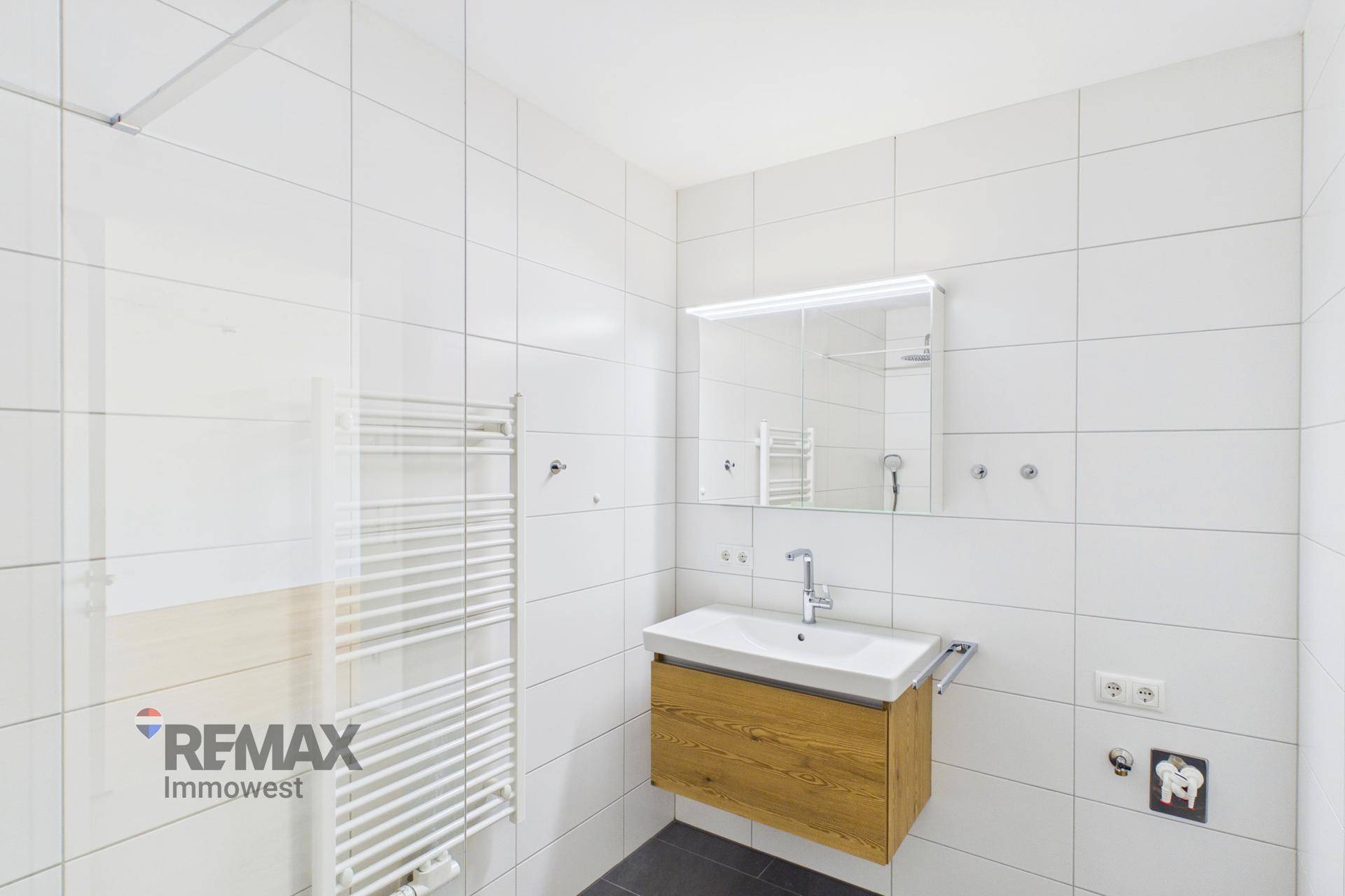 Modernes Badezimmer mit weißen Fliesen, Holzwaschtisch und beleuchtetem Spiegelschrank.
