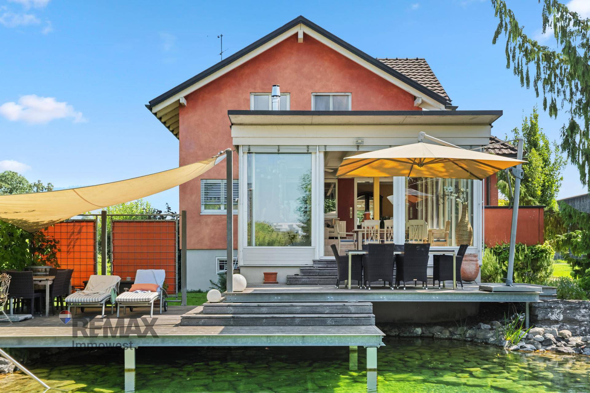 Detailansicht der Holzterrasse mit Sonnenschirm und Liegestühlen, direkt am grünen Teich gelegen.