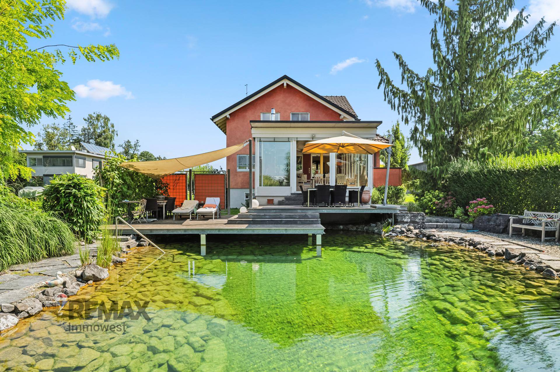 Geräumige Terrasse mit Esstisch und Liegestühlen am Teich, umgeben von grüner Natur.
