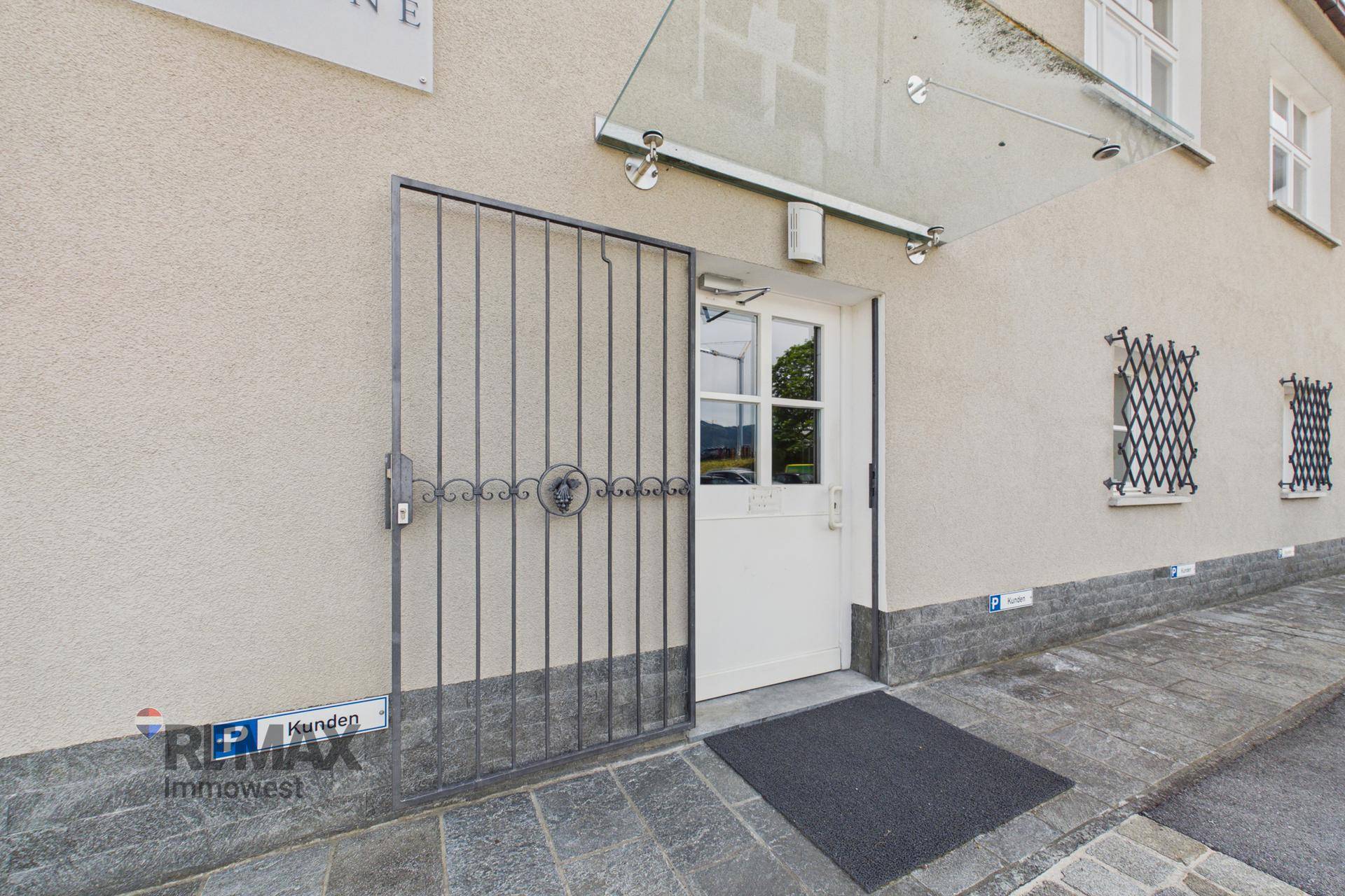 Eingangsbereich des Gebäudes mit schmiedeeisernem Tor und Glasvordach, umgeben von heller Fassade.