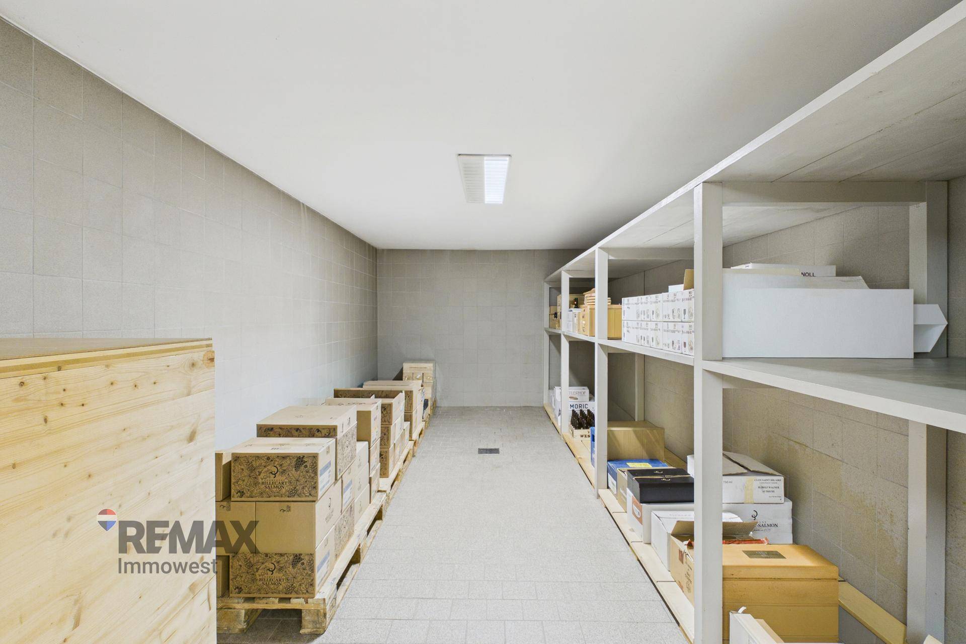Langer, schmaler Lagerraum mit Regalen an der Wand und gestapelten Kisten auf Paletten.