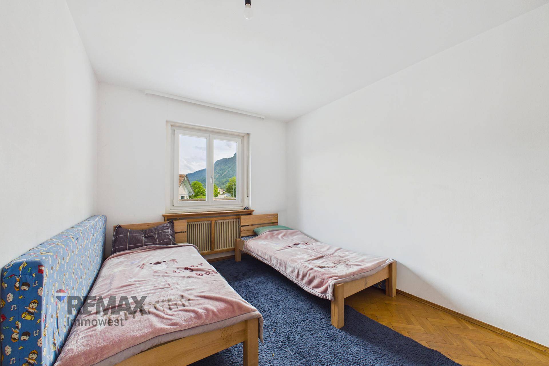 Helles Schlafzimmer mit zwei Einzelbetten, blauem Teppich und Bergblick durch das Fenster.