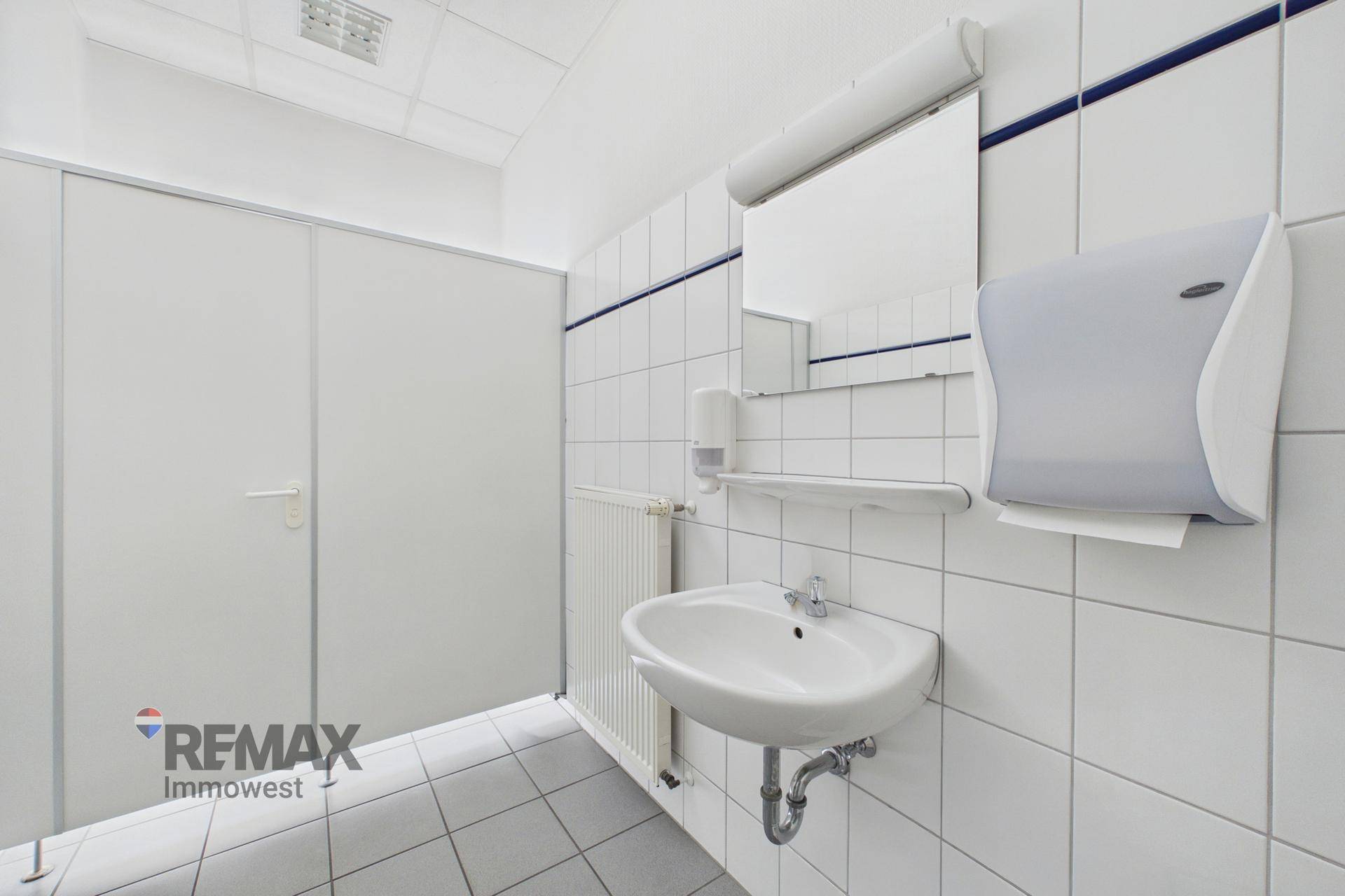Sanitärraum mit weiß gefliesten Wänden, einem Waschbecken und einem Spender für Papierhandtücher.