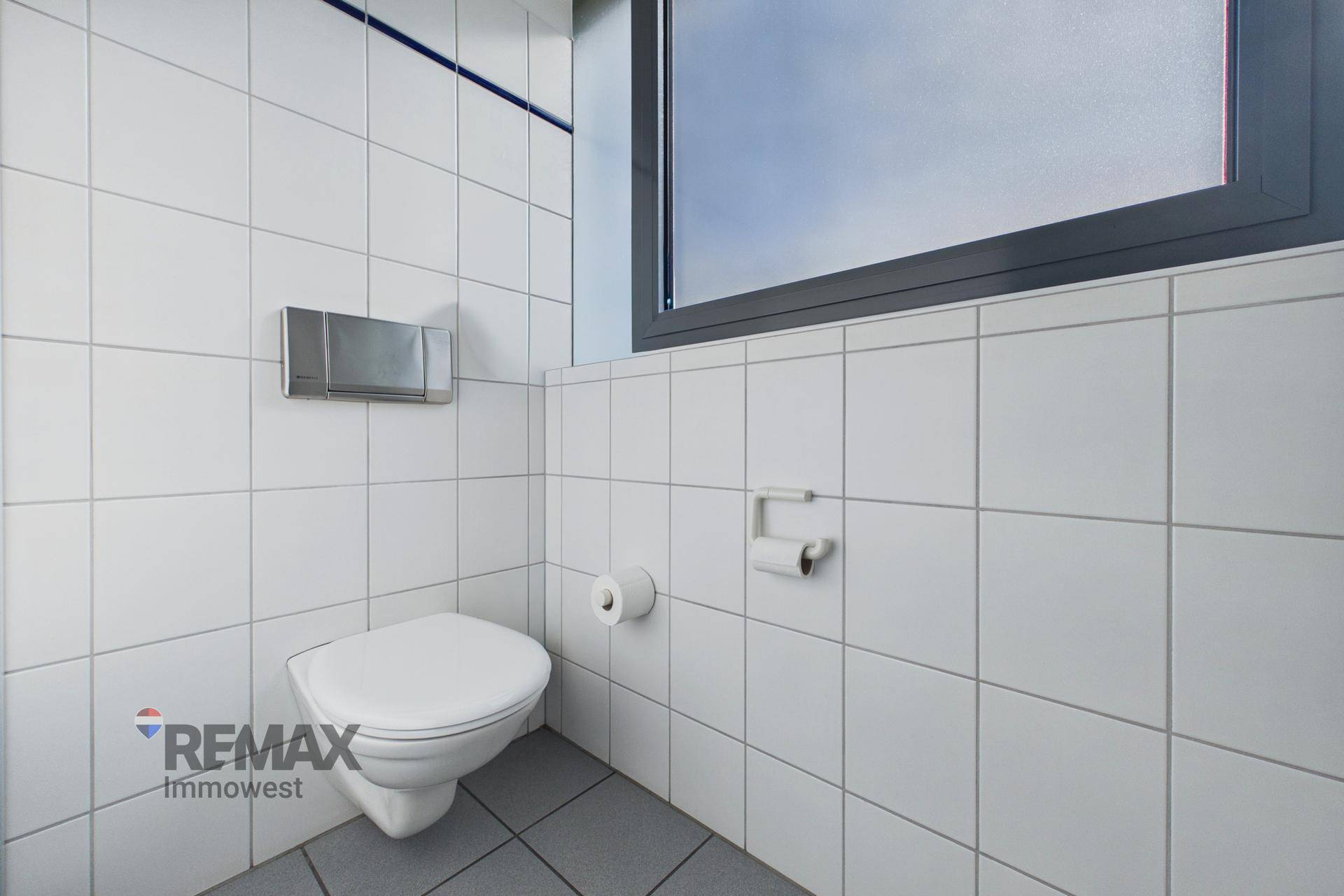 Toilettenbereich mit weiß gefliesten Wänden, einer Wandtoilette und einem Fenster.