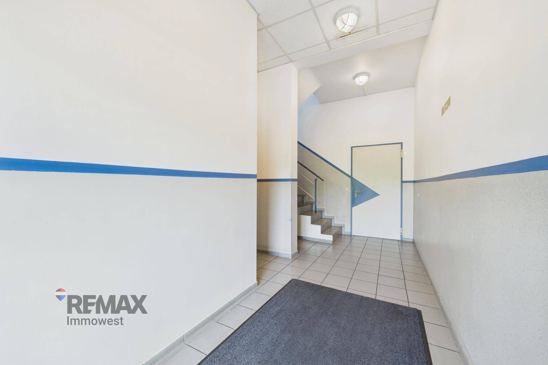 Heller Flur mit weißen Wänden, blauen Akzentstreifen und Fliesenboden, der zu einer Treppe führt.