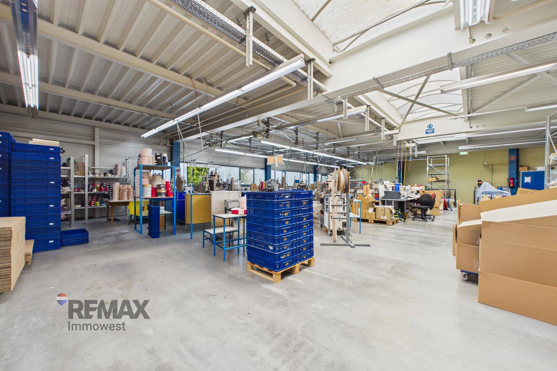 Weitläufiger Produktionsbereich mit Lagerregalen, Arbeitsplätzen und blauen Transportboxen auf dem Betonboden.