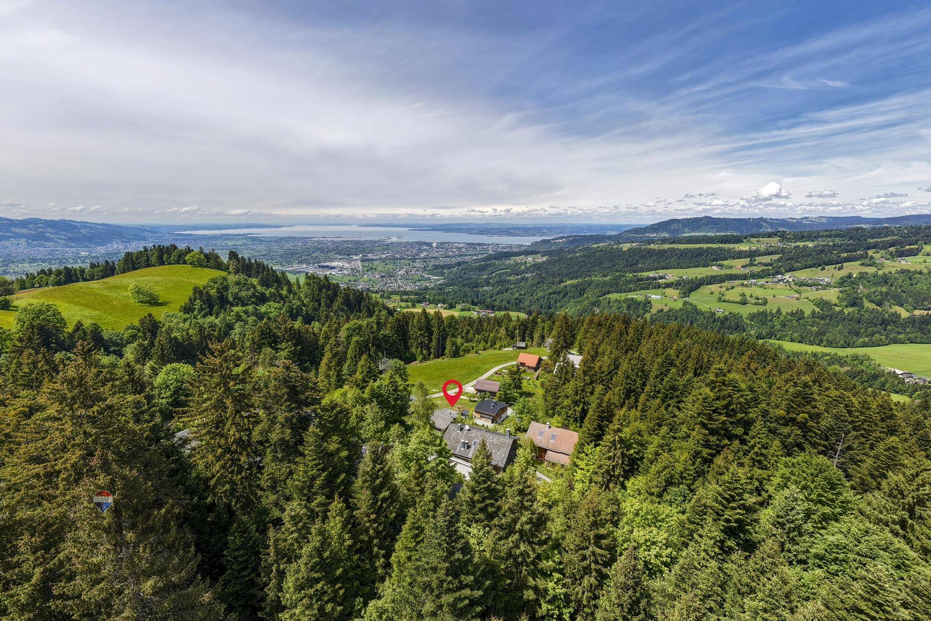 Luftaufnahme einer bewaldeten Hügellandschaft mit verstreuten Häusern und einem See in der Ferne.