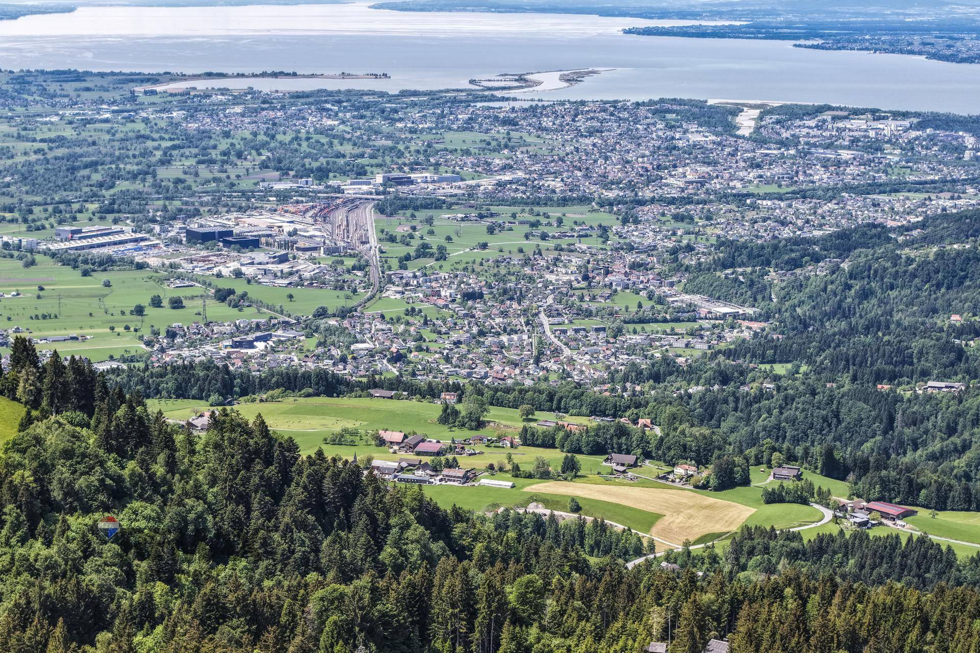 Weitwinkel-Luftaufnahme einer großen Stadt am See, umgeben von grünen Hügeln und Wäldern.