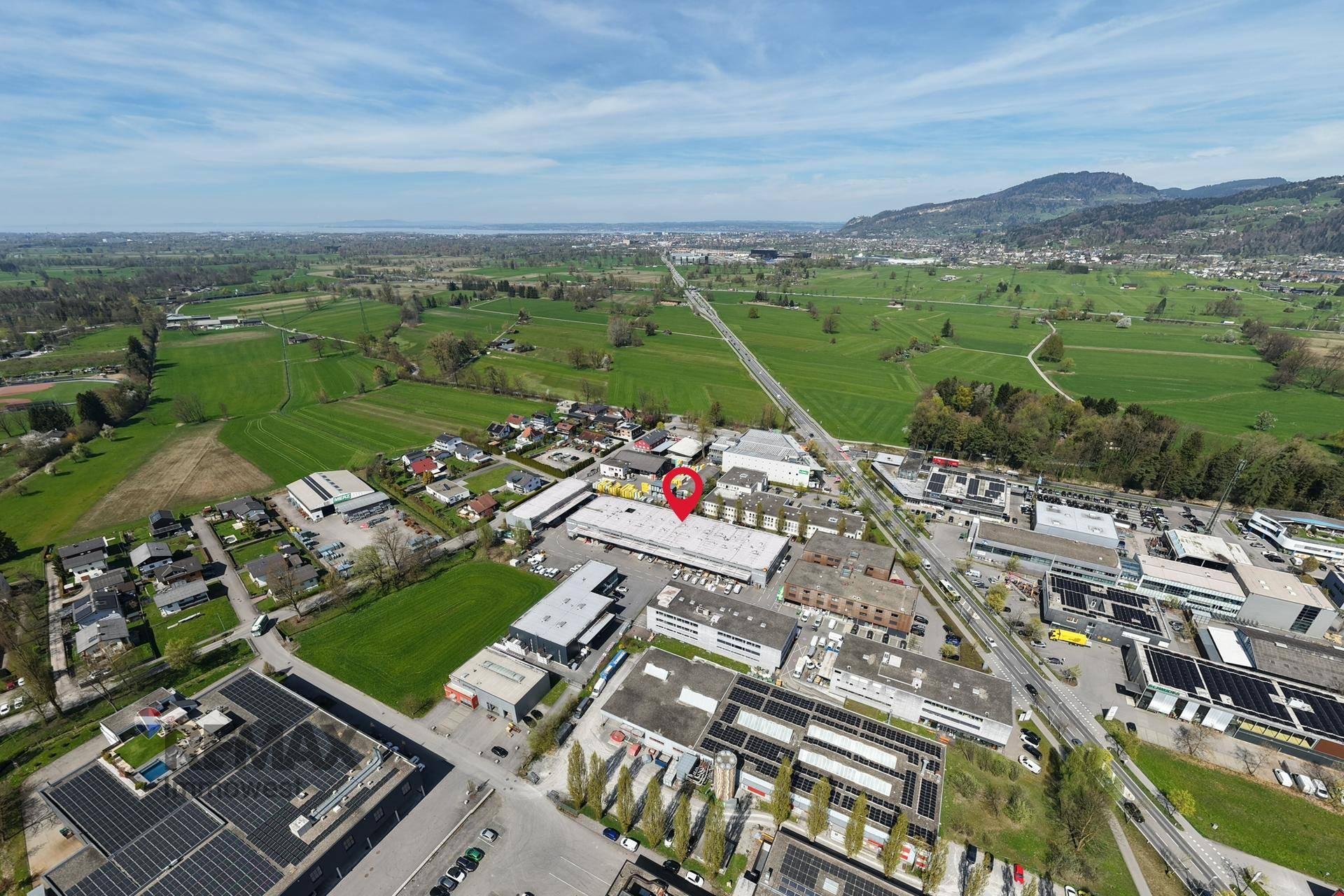 Luftaufnahme eines Gewerbegebiets mit umliegenden Feldern und Bergen im Hintergrund, Standort markiert.