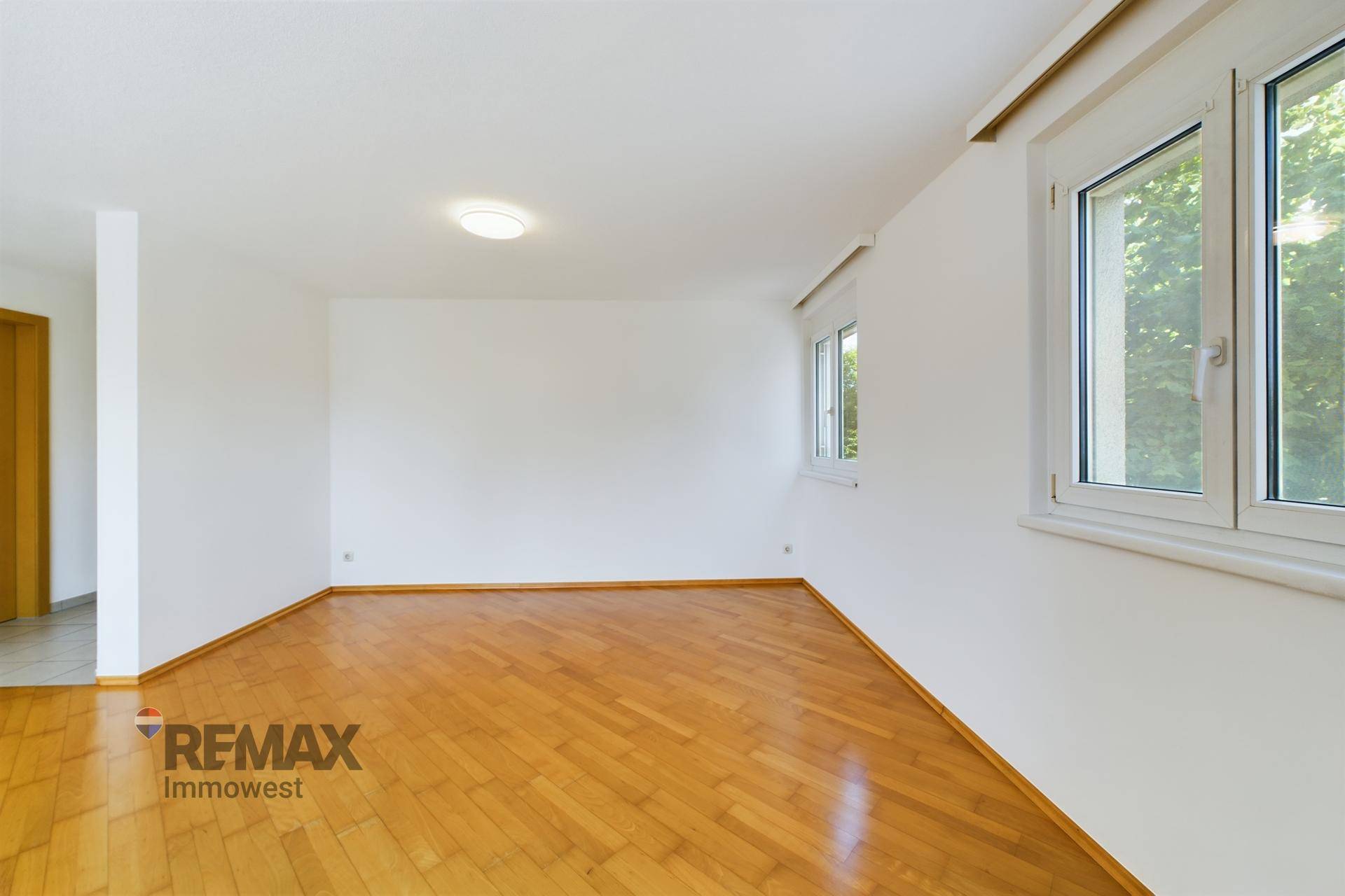 Geräumiges Wohnzimmer mit hellem Parkettboden und großen Fenstern, die viel Tageslicht hereinlassen.