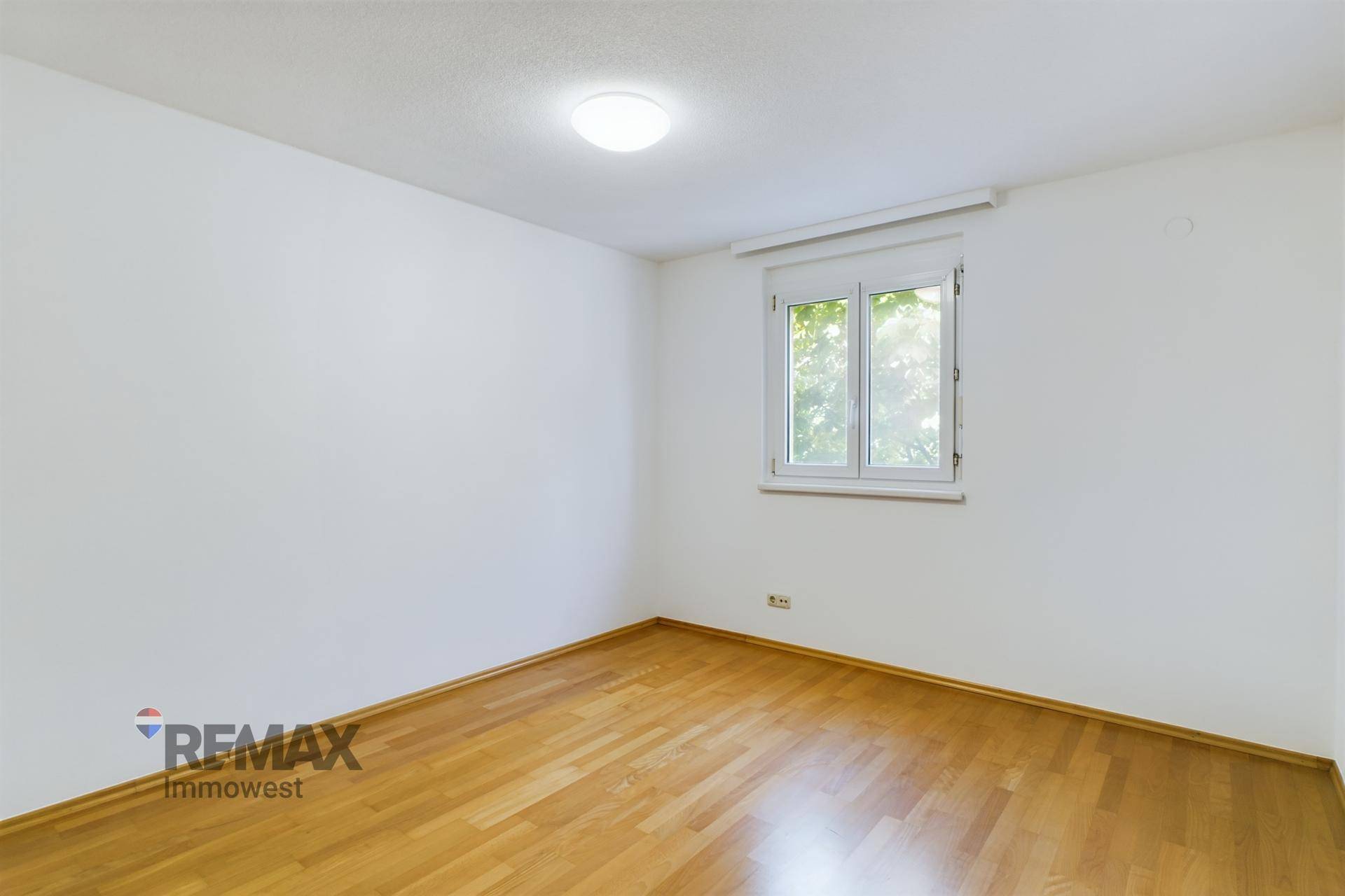 Helles Schlafzimmer mit Parkettboden und einem Fenster mit Blick ins Grüne.