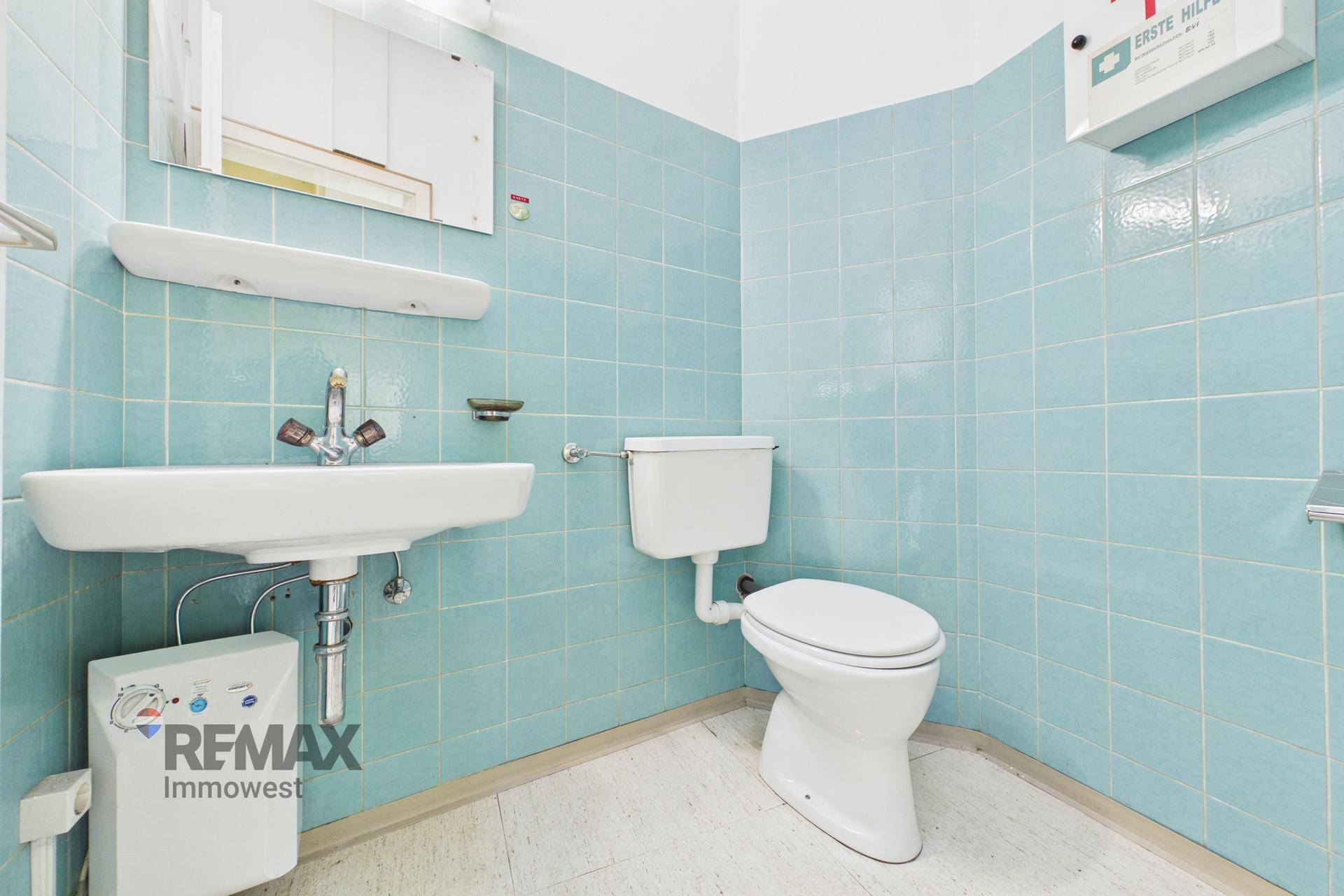 Einfache Toilette mit hellblauen Fliesen, Waschbecken und einem Erste-Hilfe-Kasten an der Wand.
