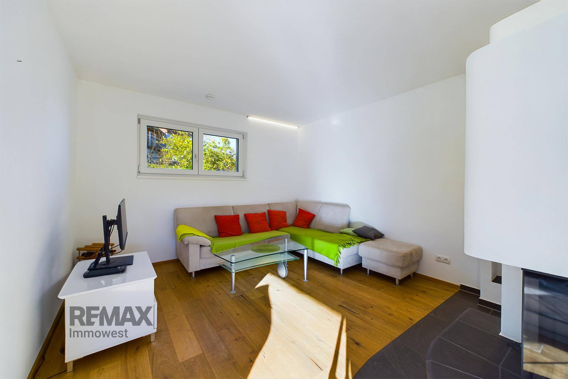 Heller Wohnbereich mit Sofa, Holzboden und Fenster mit Blick auf die grüne Umgebung.