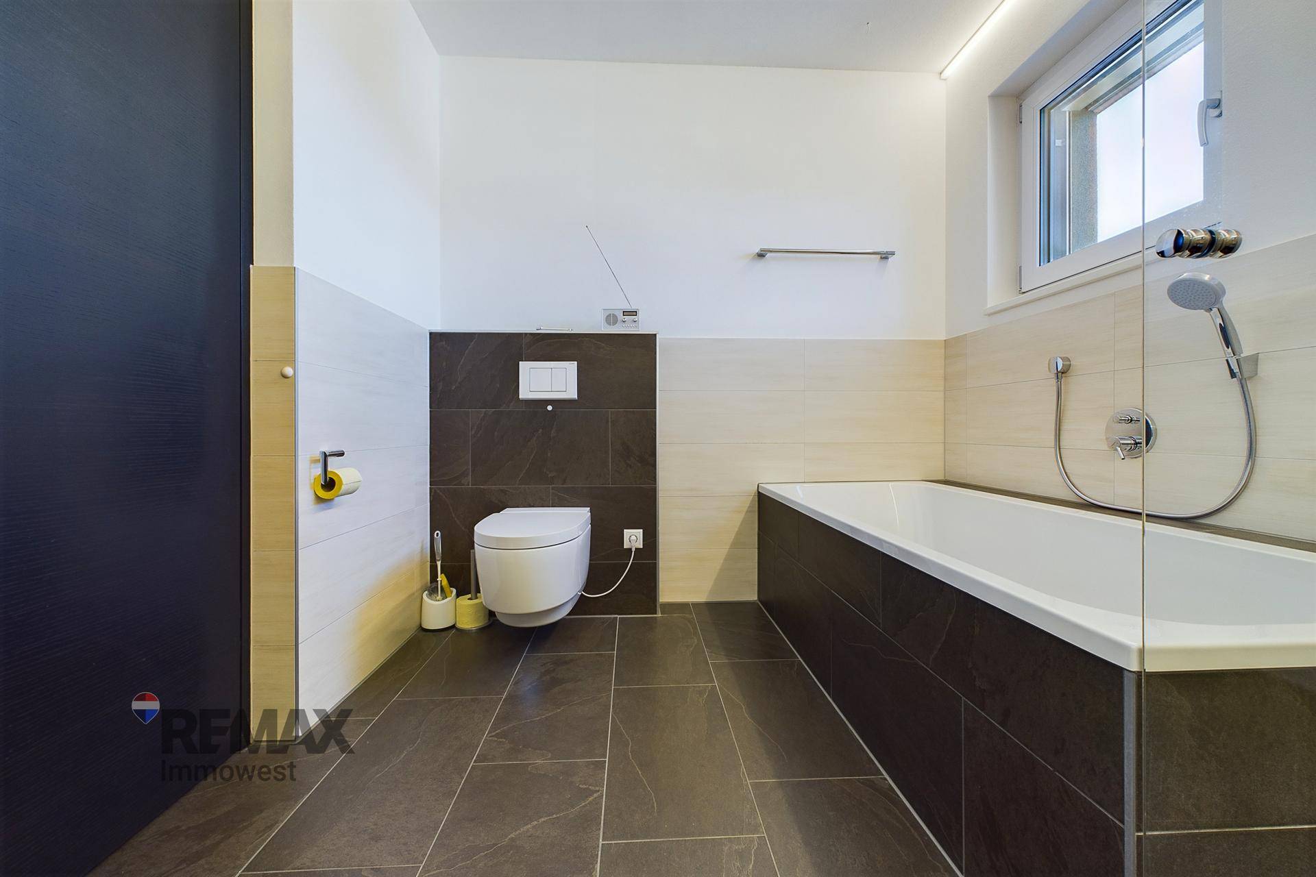 Helles Badezimmer mit Badewanne, wandhängendem WC und Fenster für Tageslicht.