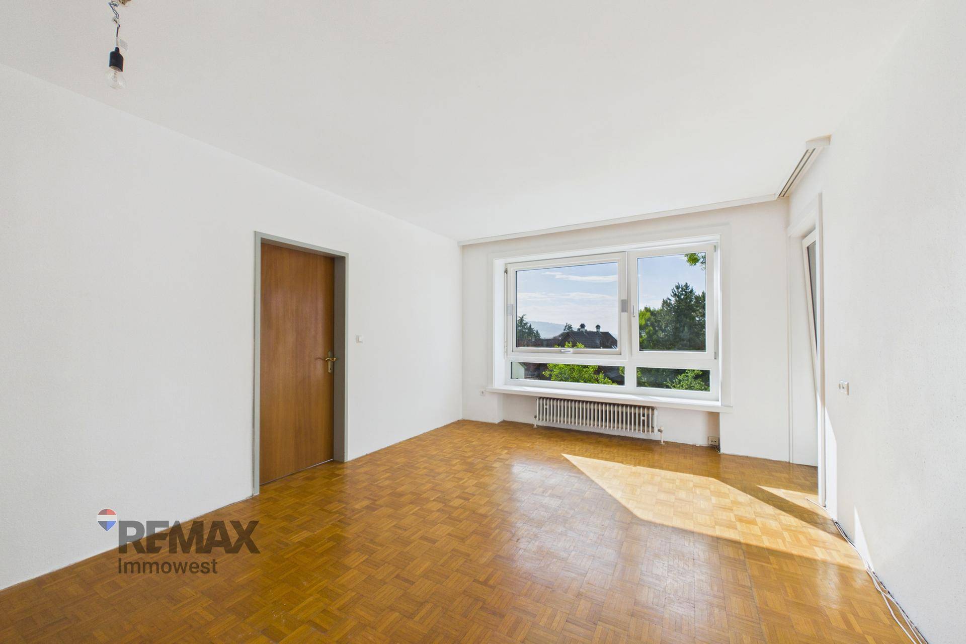 Geräumiges Wohnzimmer mit hellem Parkettboden und viel Tageslicht durch das große Fenster.