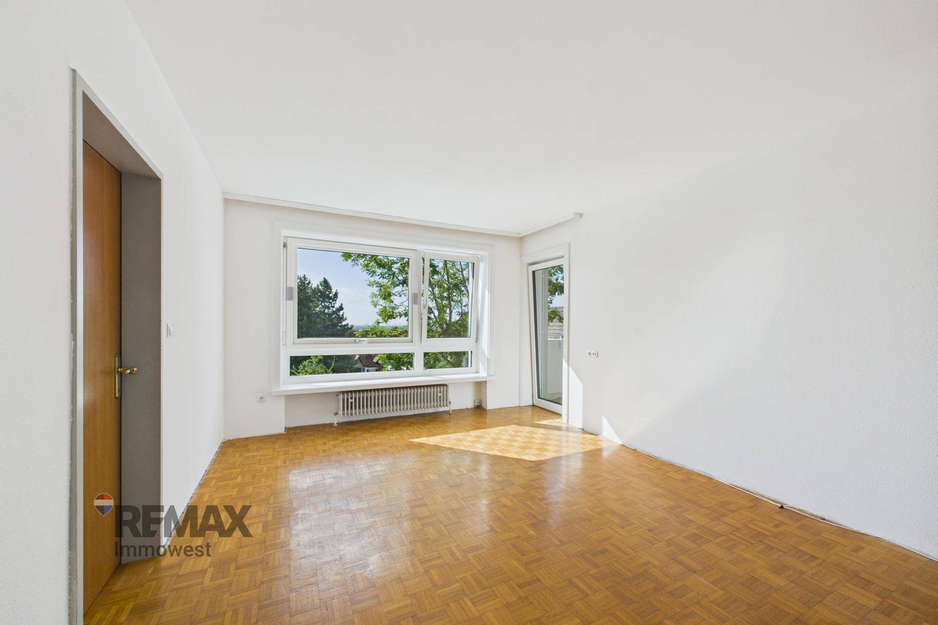 Helles, leeres Wohnzimmer mit Fischgrätparkett und großem Fenster mit Blick ins Grüne.