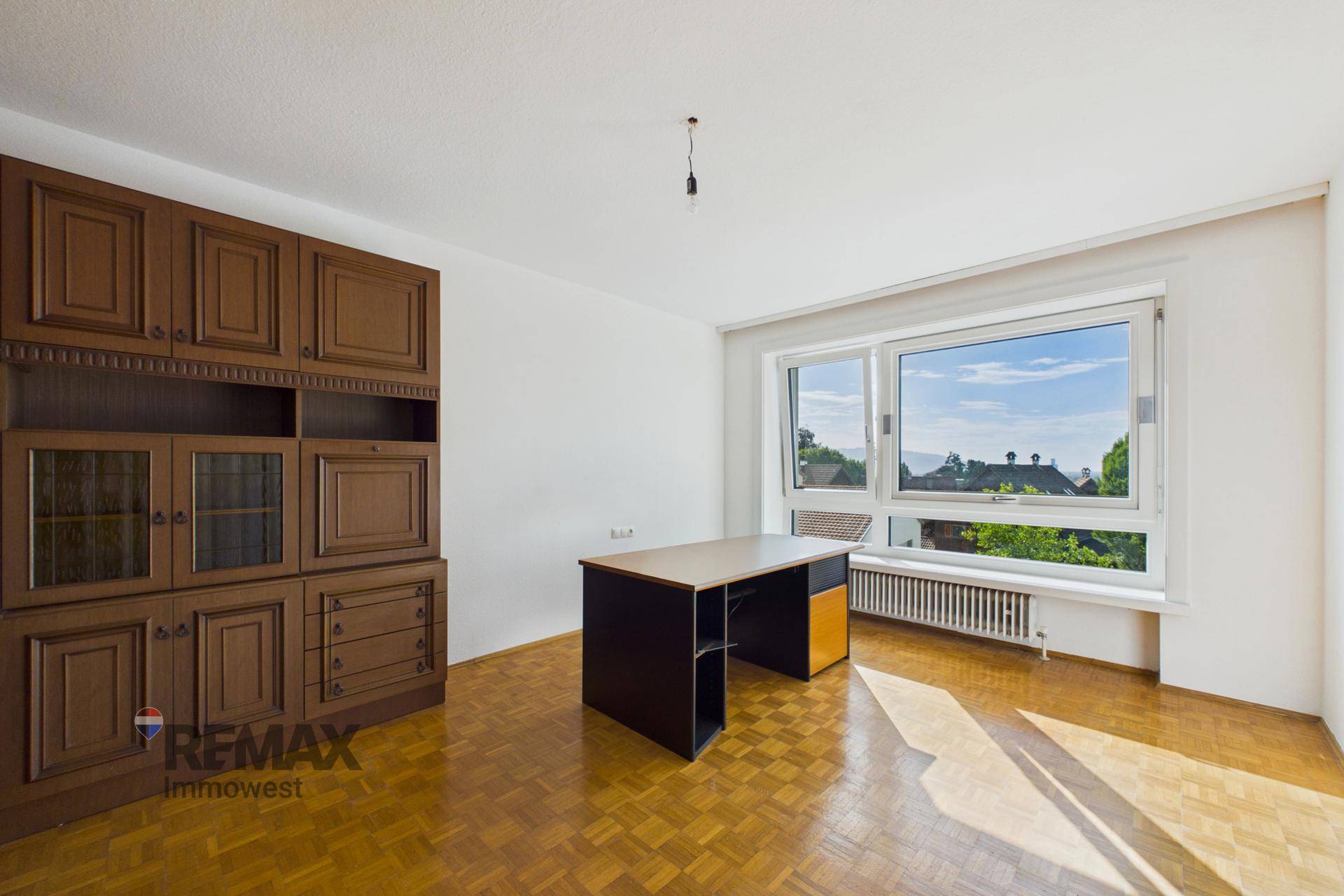 Geräumiges Zimmer mit Holzschrank, Schreibtisch und Blick ins Grüne durch das große Fenster.