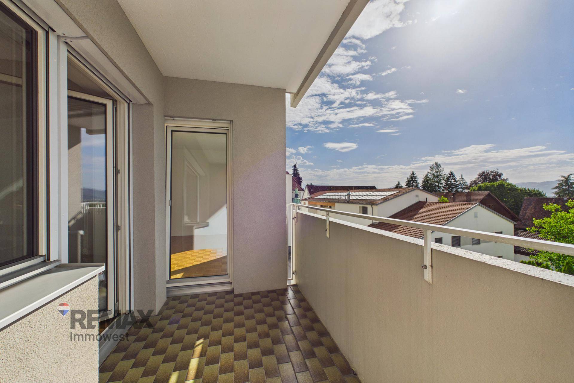 Sonniger Balkon mit Fliesenboden und Zugang zum Innenbereich, umgeben von Grün.