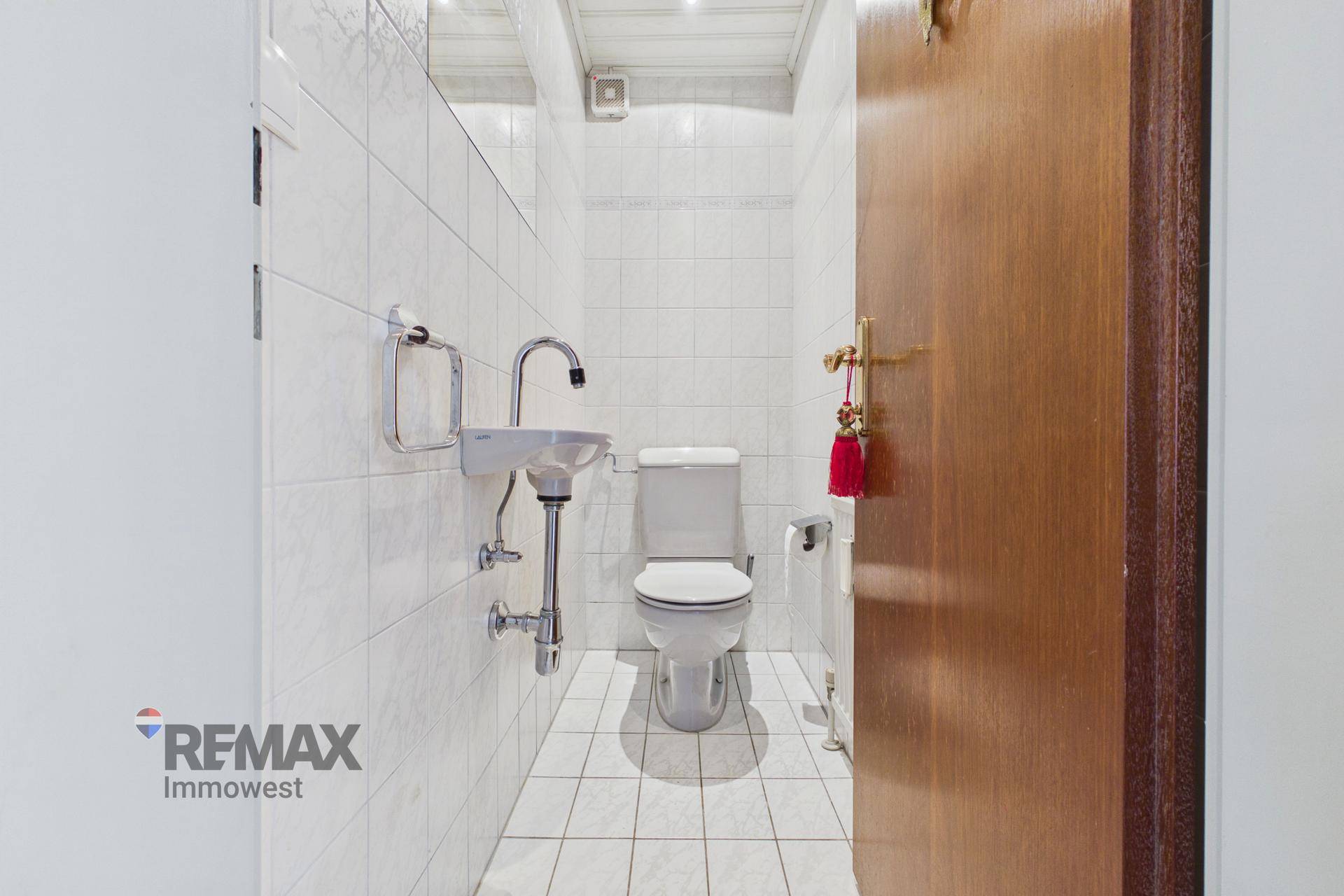 Separates WC mit weißen Fliesen, Toilette und kleinem Handwaschbecken.