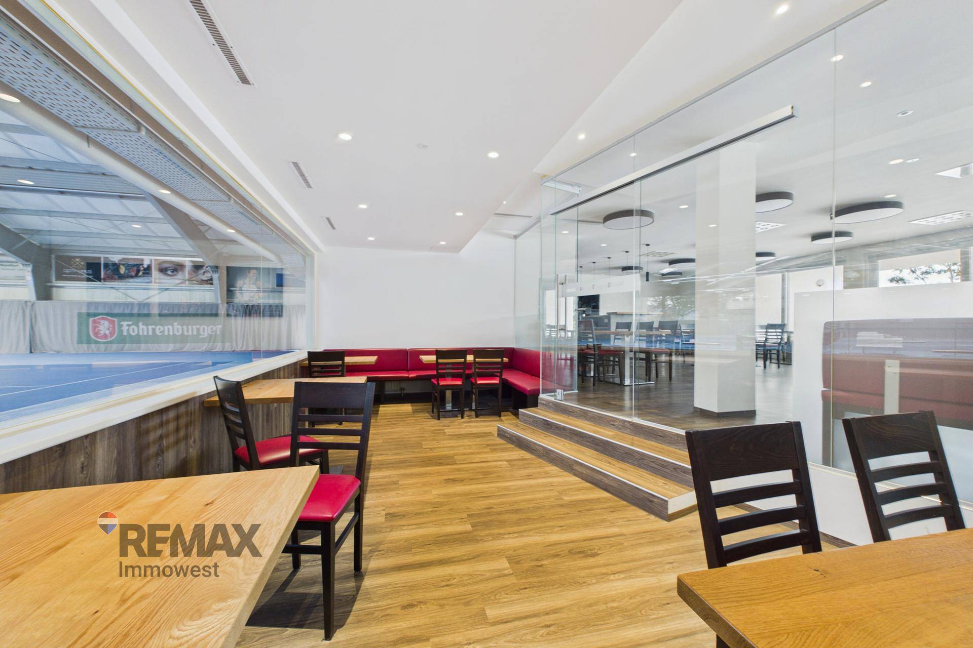 Gemütlicher Restaurantbereich mit roten Sitzbänken und Blick durch eine große Glasfront auf eine Sportanlage.