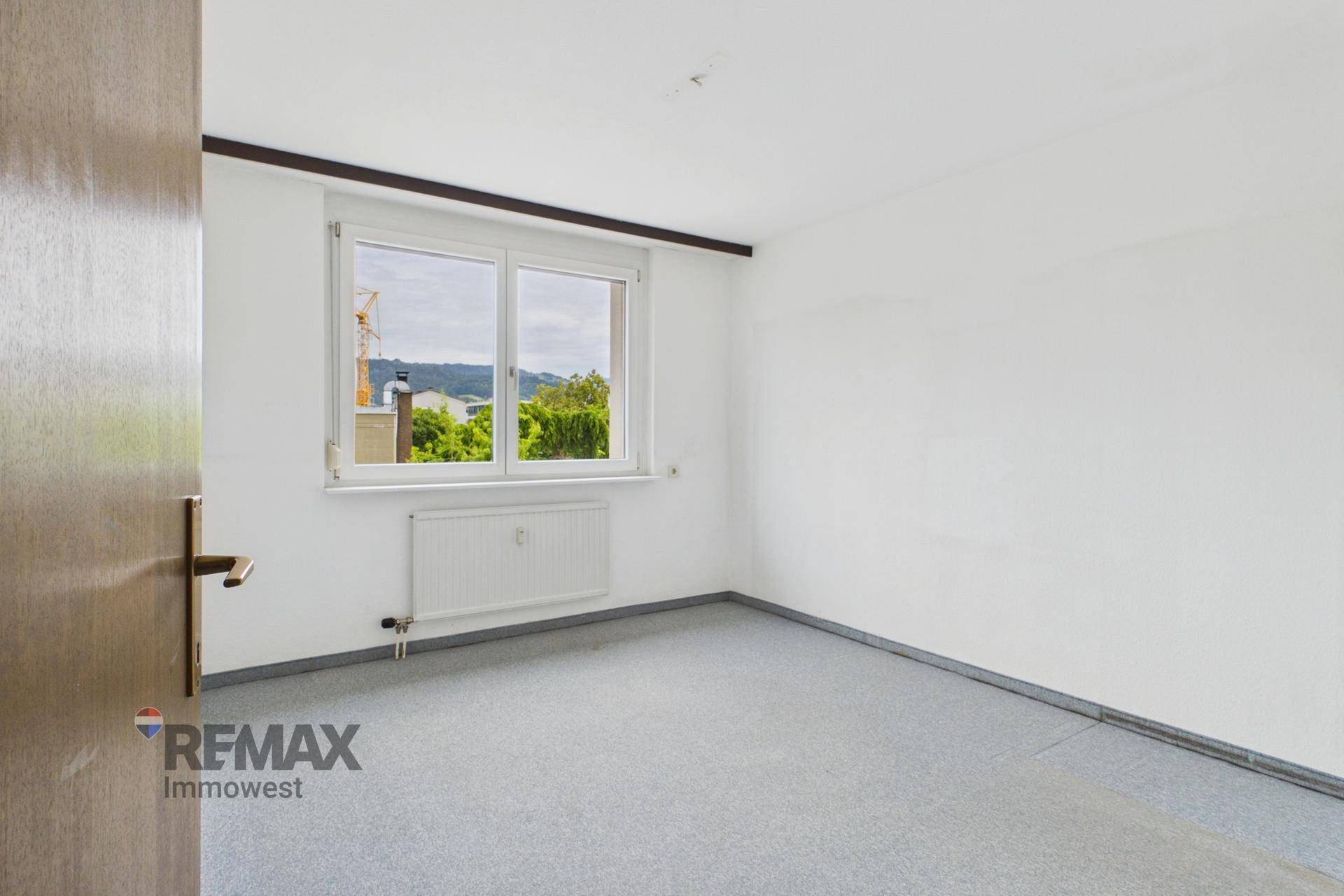 Helles Zimmer mit Teppichboden, Heizkörper und Fenster mit Blick ins Grüne.