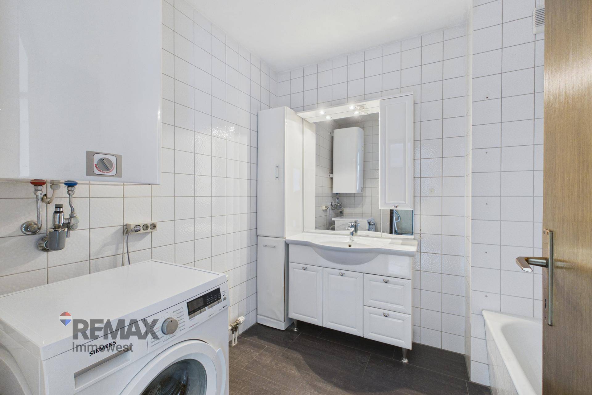 Badezimmer mit weißen Fliesen, Waschbeckenunterschrank, Waschmaschine und Warmwasserboiler.