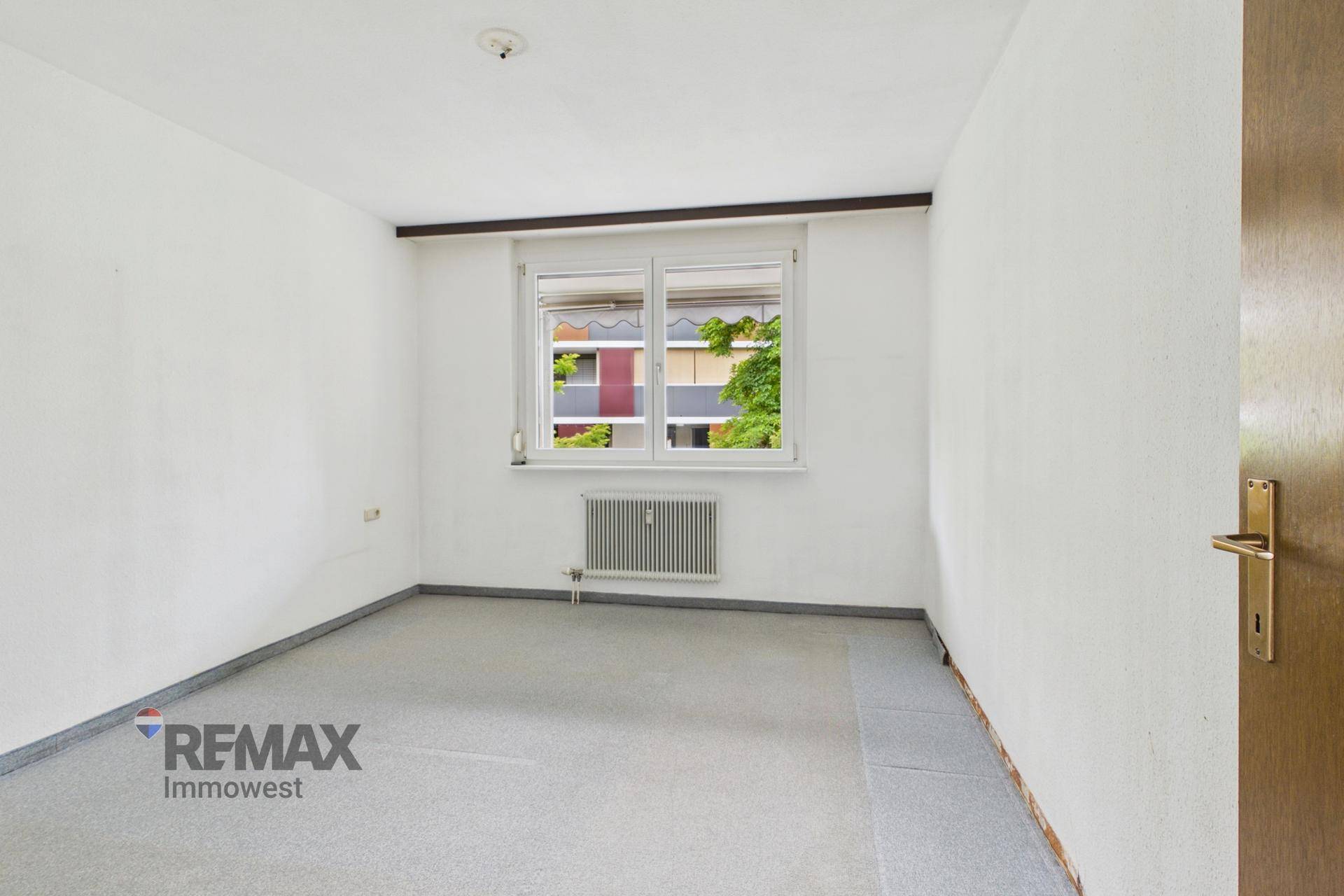Leeres Zimmer mit Teppichboden, Heizkörper und Fenster mit Blick auf die Umgebung.