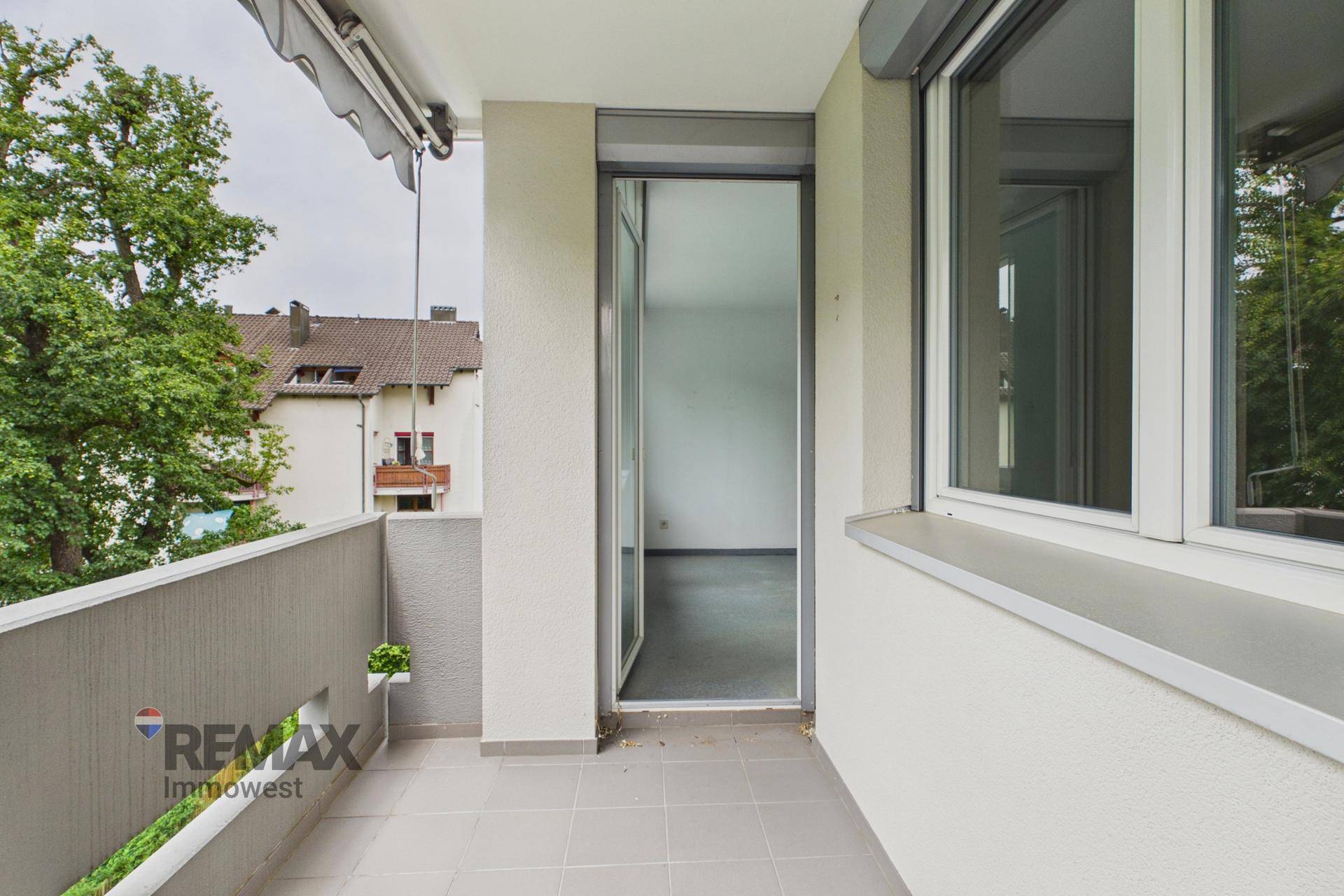 Balkon mit grauen Fliesen, Markise und Blick auf Bäume und Nachbargebäude.
