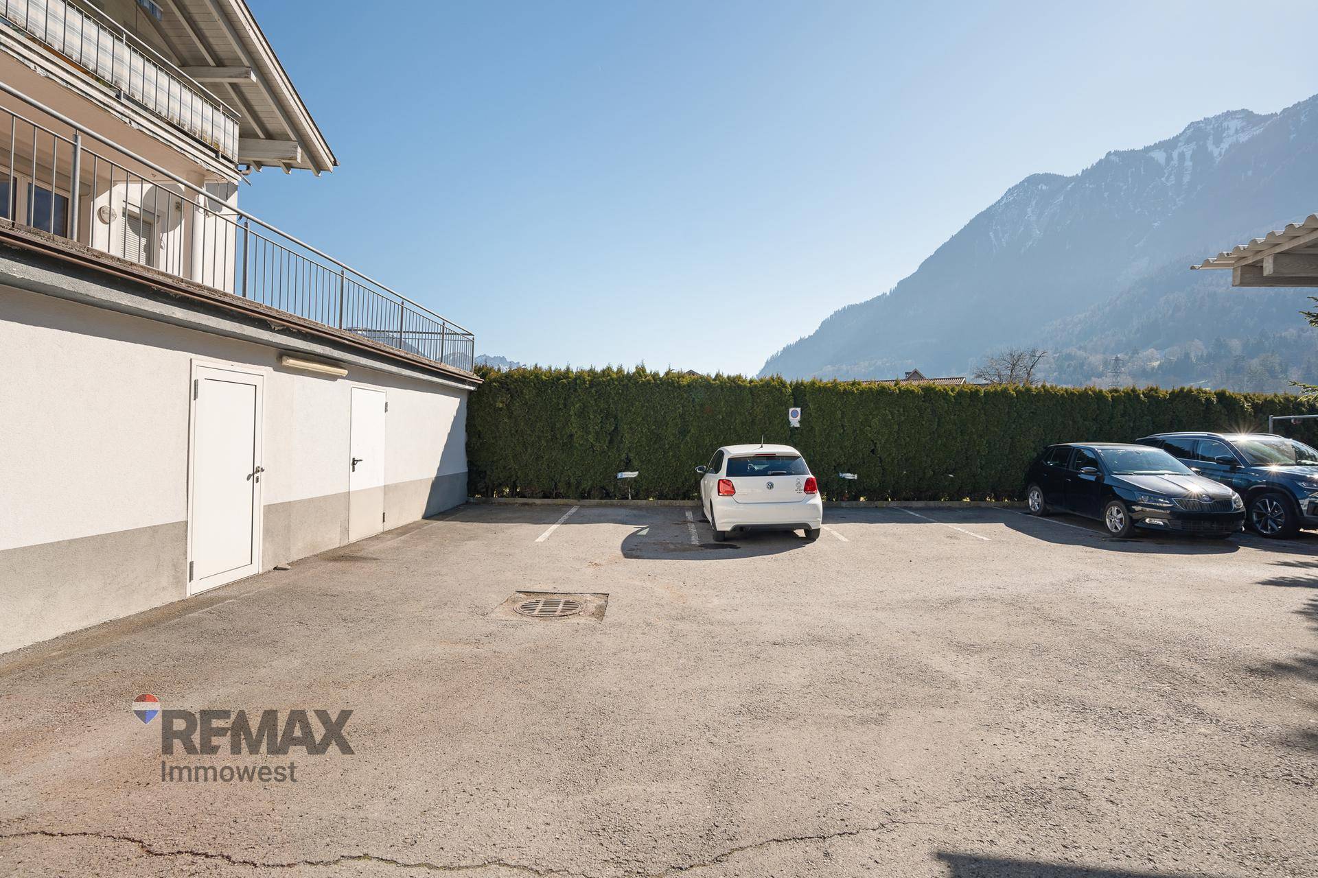 Außenparkplatz mit Hecke und Blick auf die umliegende Berglandschaft unter blauem Himmel.