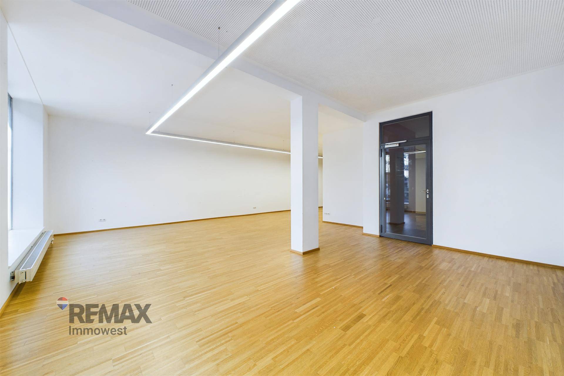 Geräumiger, heller Büroraum mit Holzparkettboden, weißen Wänden und moderner Deckenbeleuchtung.