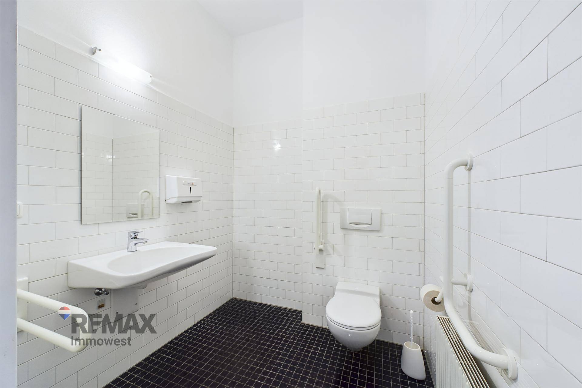 Modernes, barrierefreies Badezimmer mit weißen Fliesen, dunklem Boden und Haltegriffen an der Toilette und am Waschbecken.