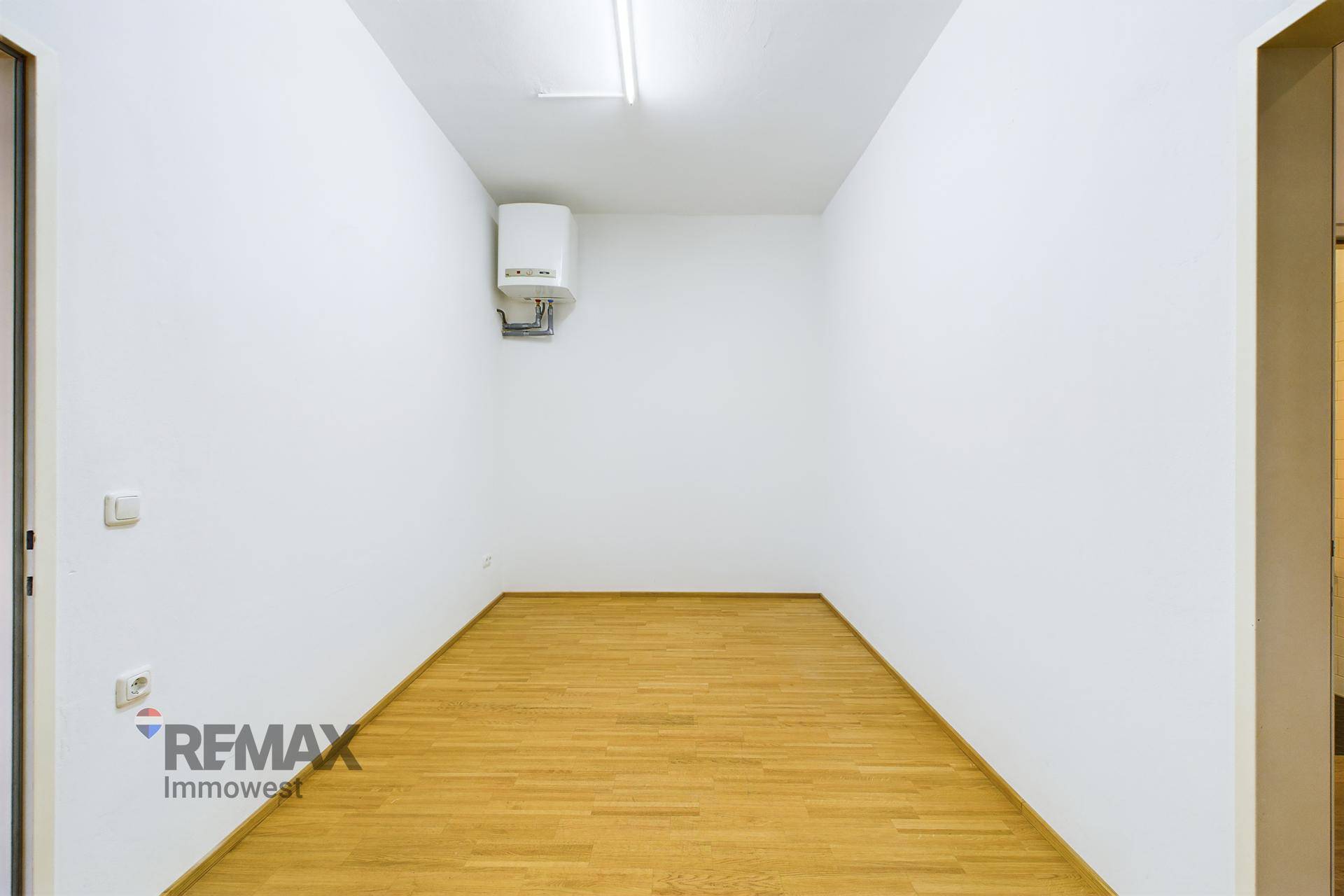 Kleiner, leerer Raum mit weiß gestrichenen Wänden, Holzboden und einem Warmwasserbereiter an der Wand.
