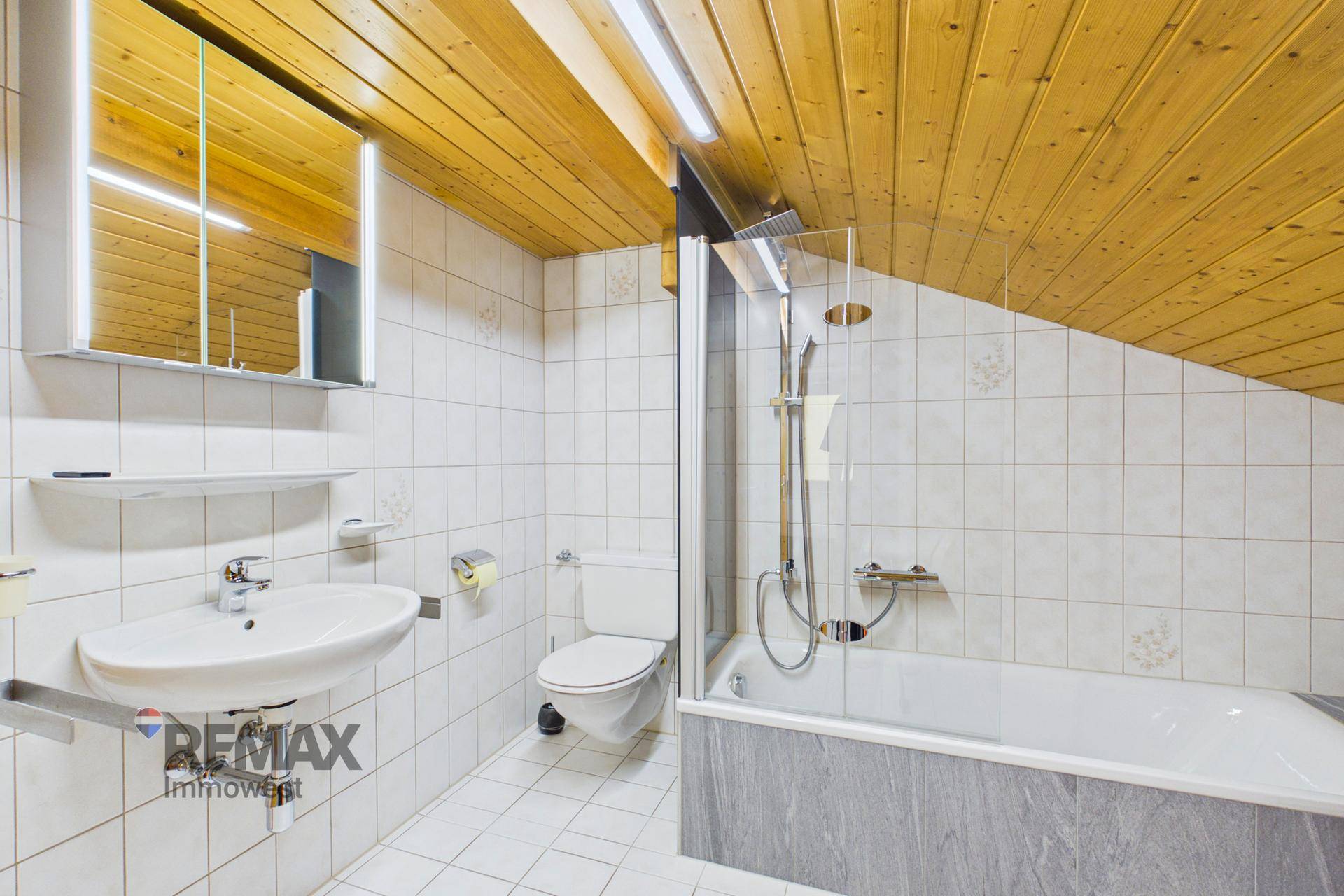 Modernisiertes Badezimmer mit weißen Fliesen, Badewanne und Dusche unter Holzdecke.