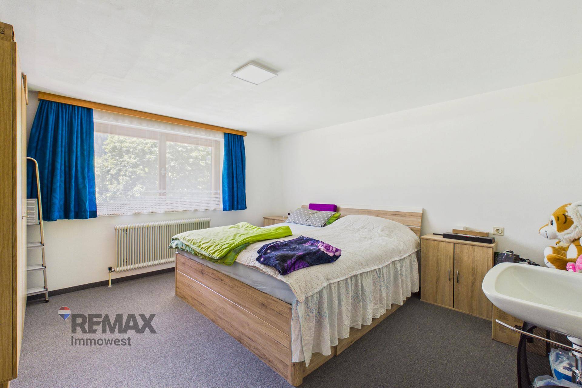 Helles Schlafzimmer mit weißen Wänden, Doppelbett, Waschbecken und großem Fenster.
