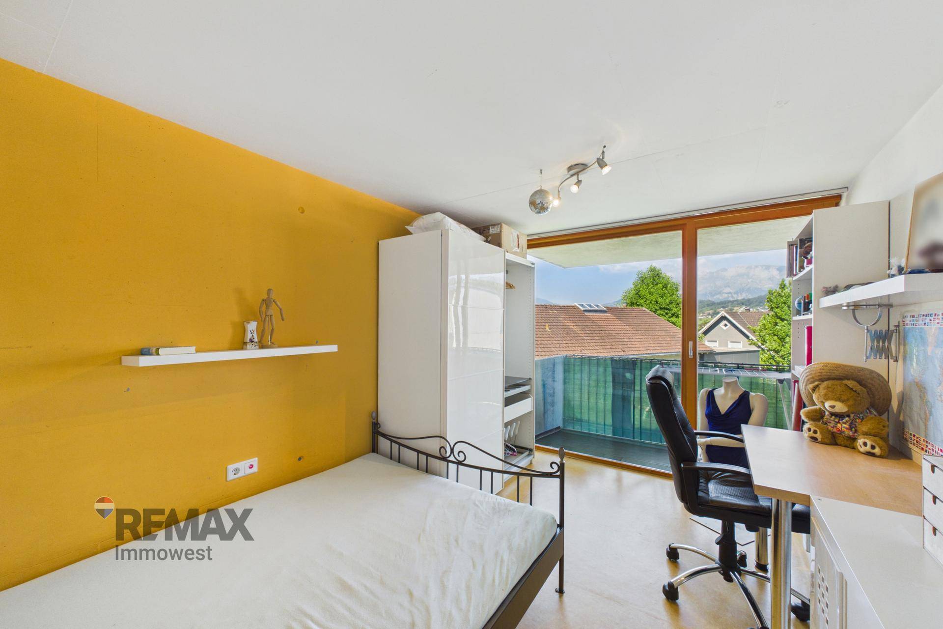 Schlafzimmer mit gelber Wand, Einzelbett, Schreibtisch und großem Fenster mit Balkonzugang.
