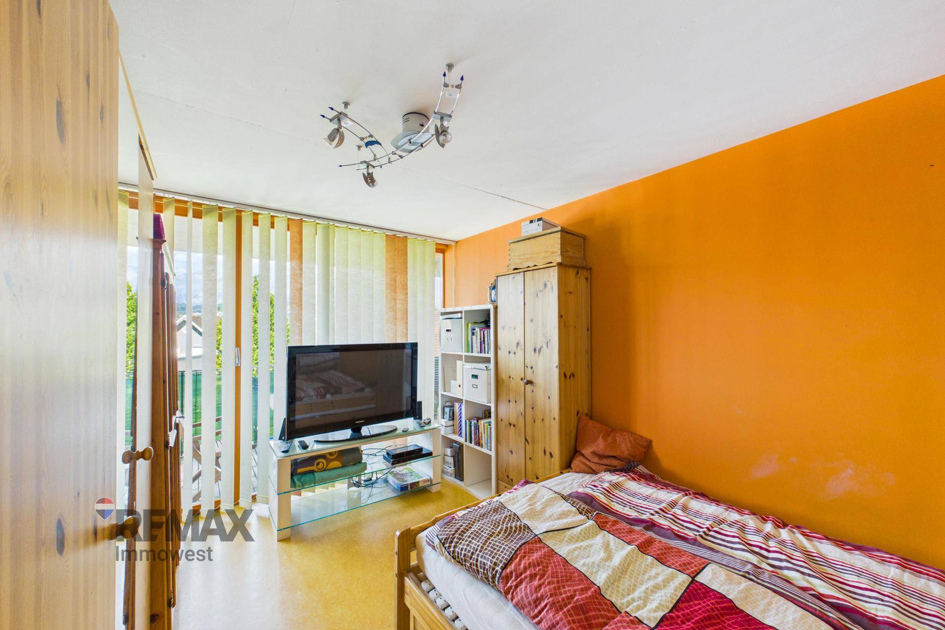Schlafzimmer mit orangefarbener Akzentwand, Einzelbett und Holzschrank, bietet Zugang nach draußen.