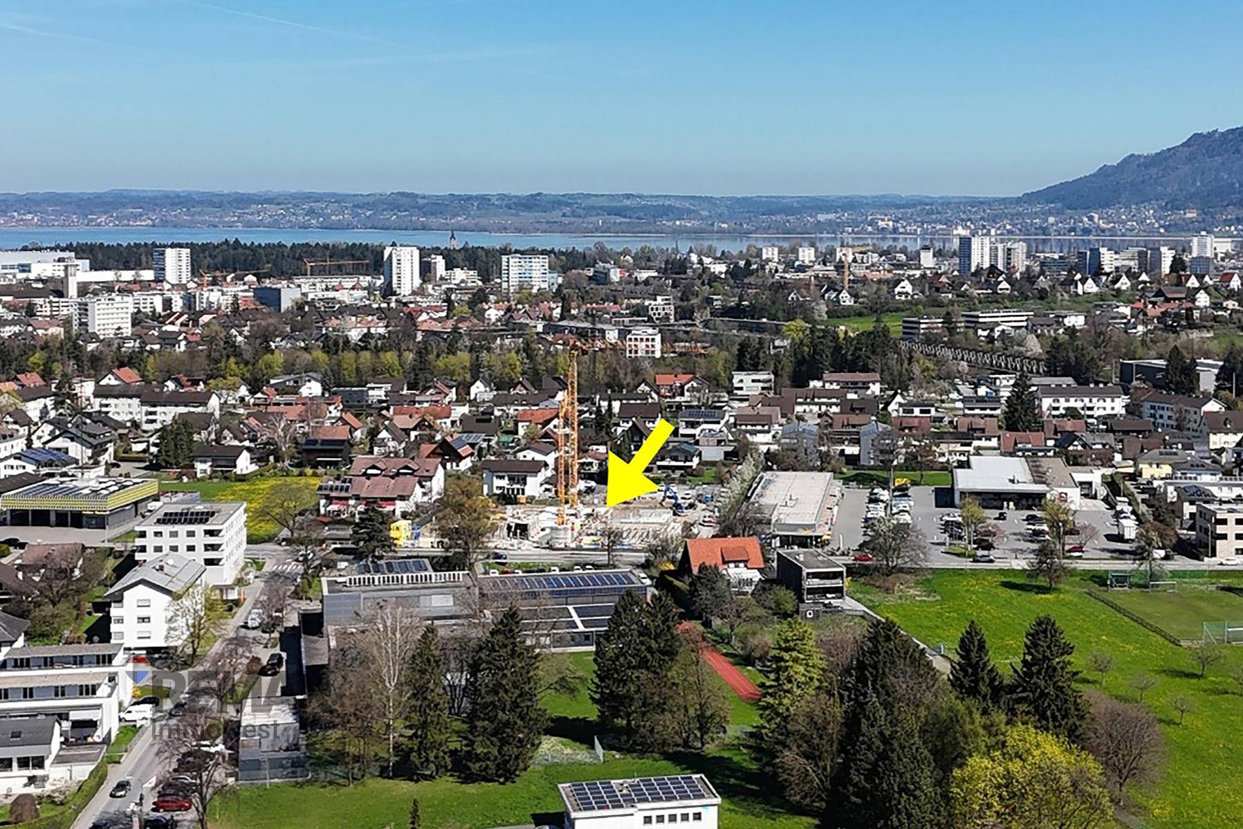 Luftaufnahme einer Stadt mit einer Baustelle, markiert durch einen gelben Pfeil, und See- sowie Bergblick.