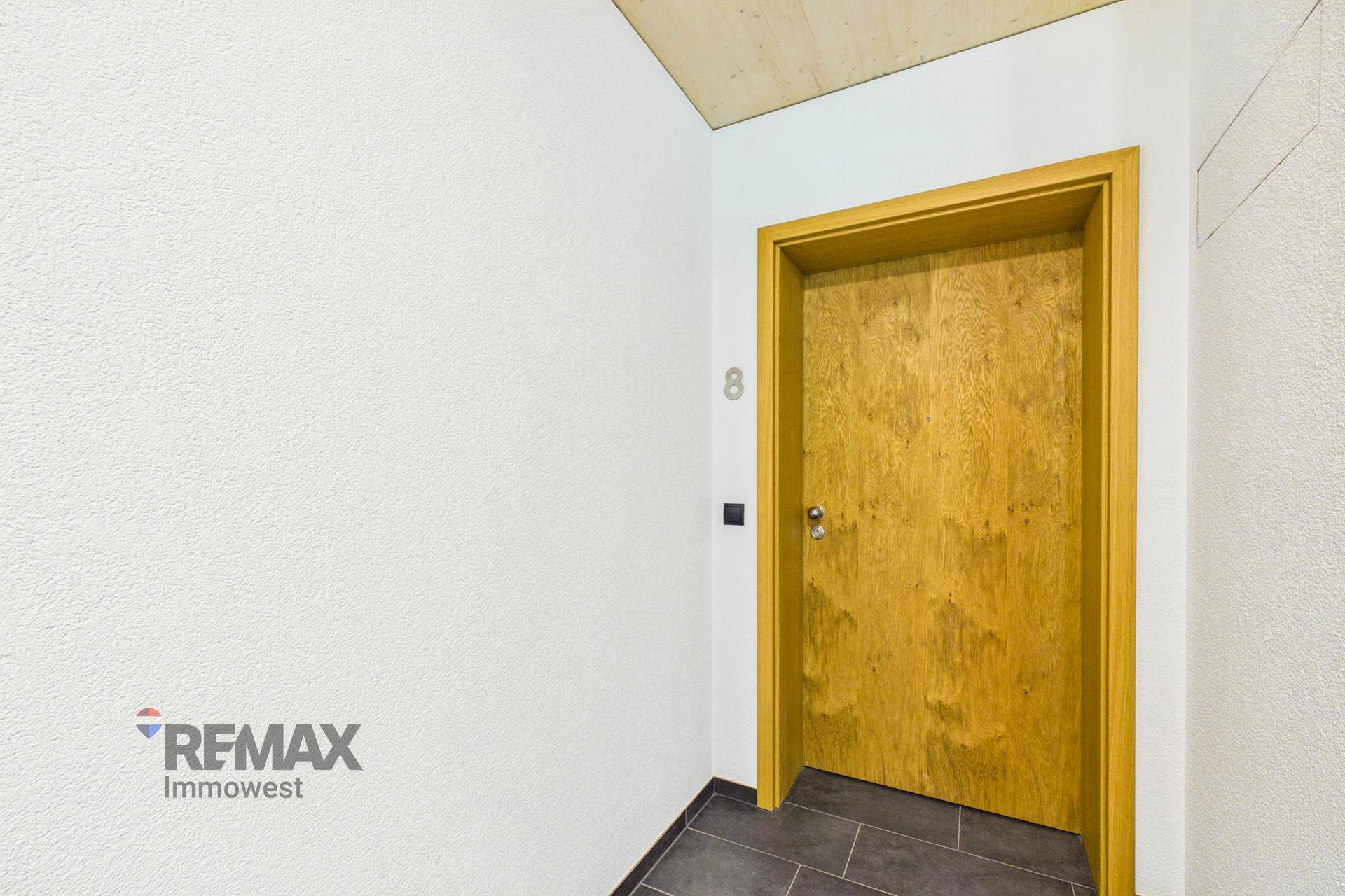 Heller Eingangsbereich mit Holztür und dunklen Fliesen, der zu einer Wohnung führt.
