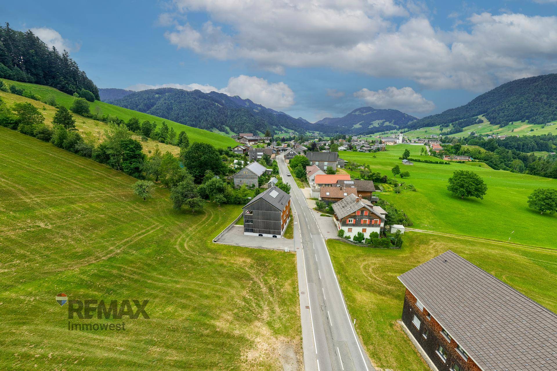 Weitläufige Luftaufnahme eines Dorfes in einem Tal, umgeben von grünen Wiesen und Bergen.