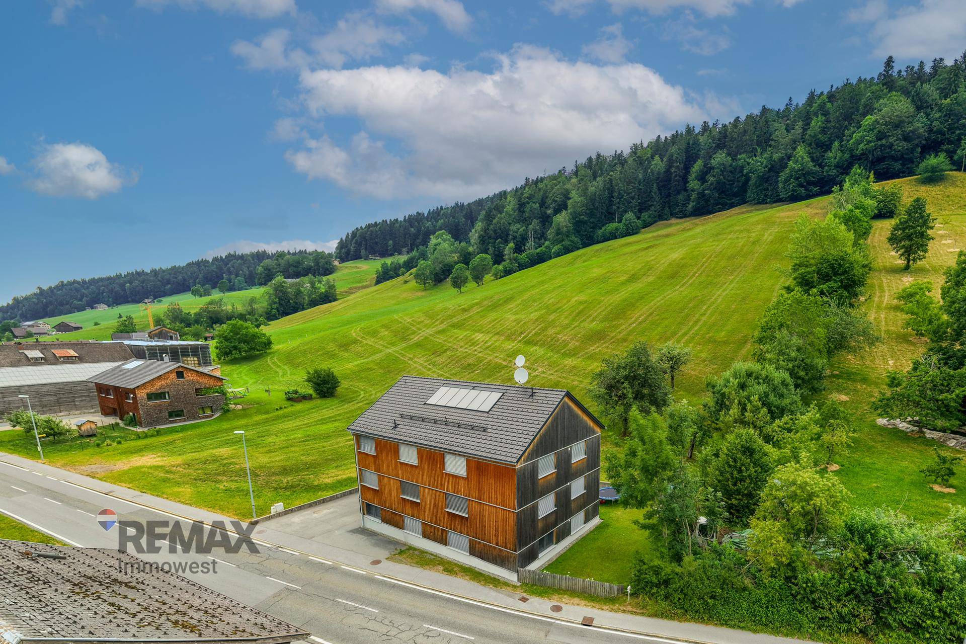 Vogelperspektive eines Holzhauses an einer Straße, umgeben von grünen Hügeln und Wäldern.