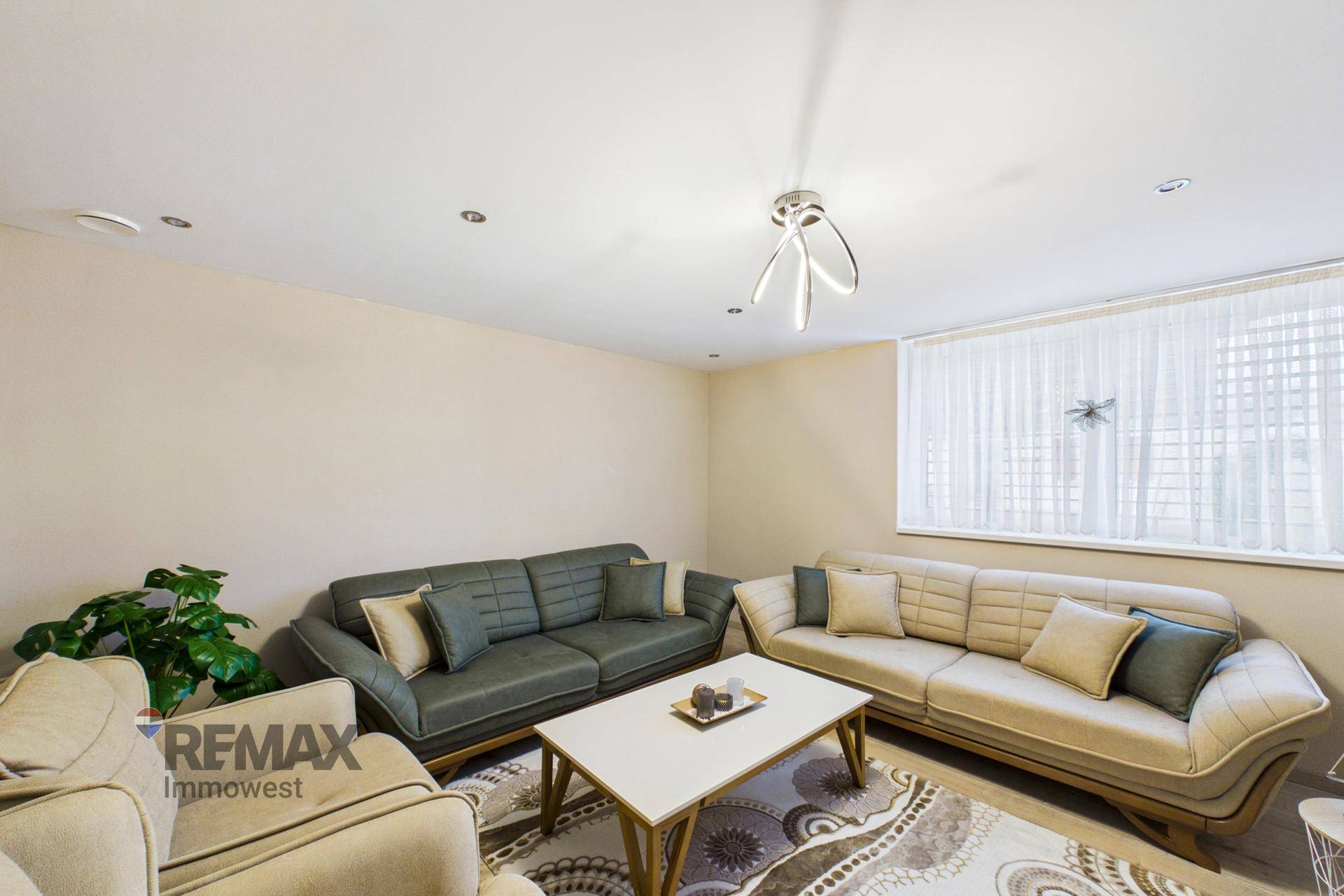 Geräumiges Wohnzimmer mit beigen Sofas, Teppich und großen Fenstern mit Vorhängen.