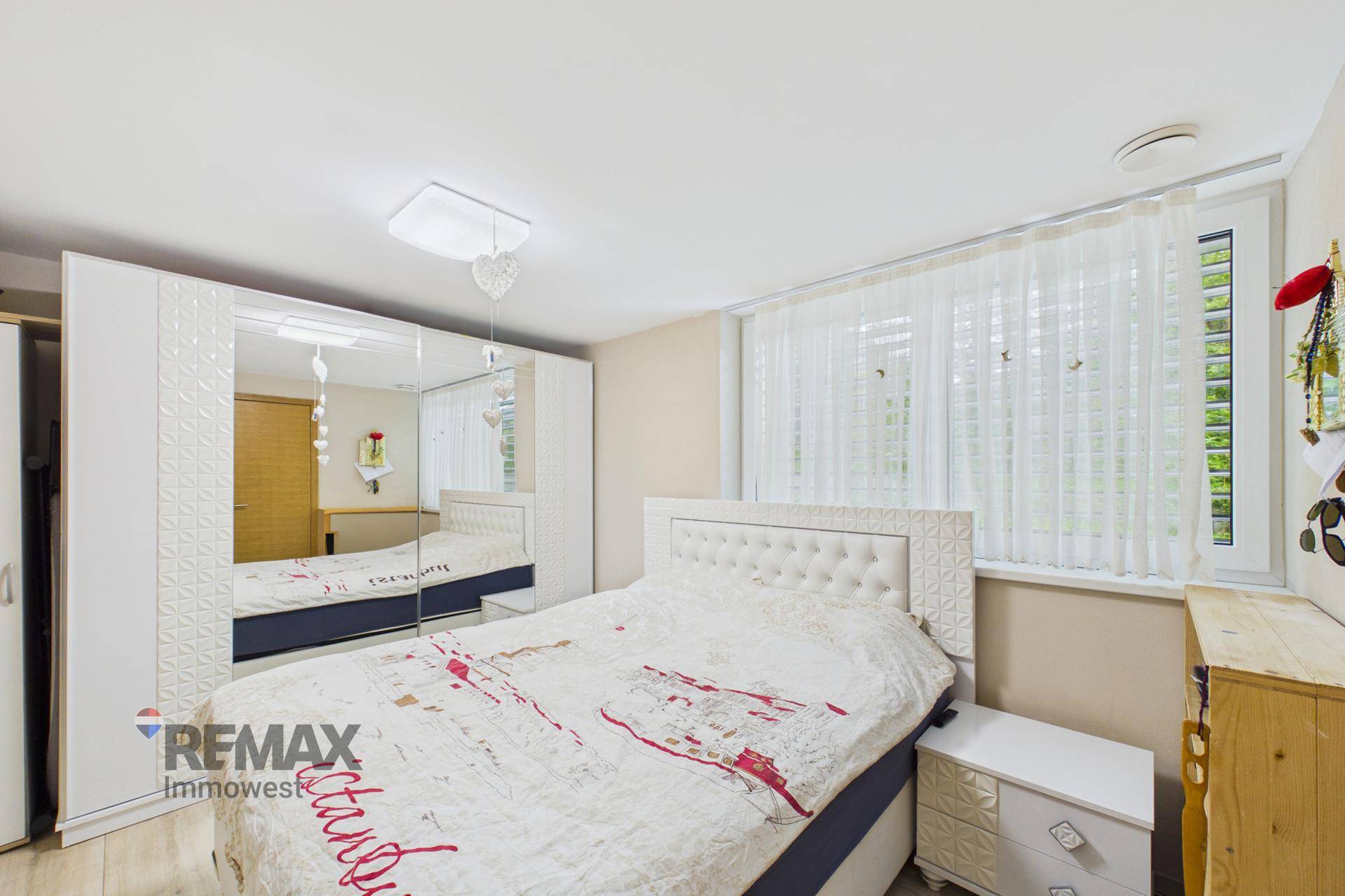 Schlafzimmer mit großem Spiegelkleiderschrank, Doppelbett und Fenster mit Vorhängen.