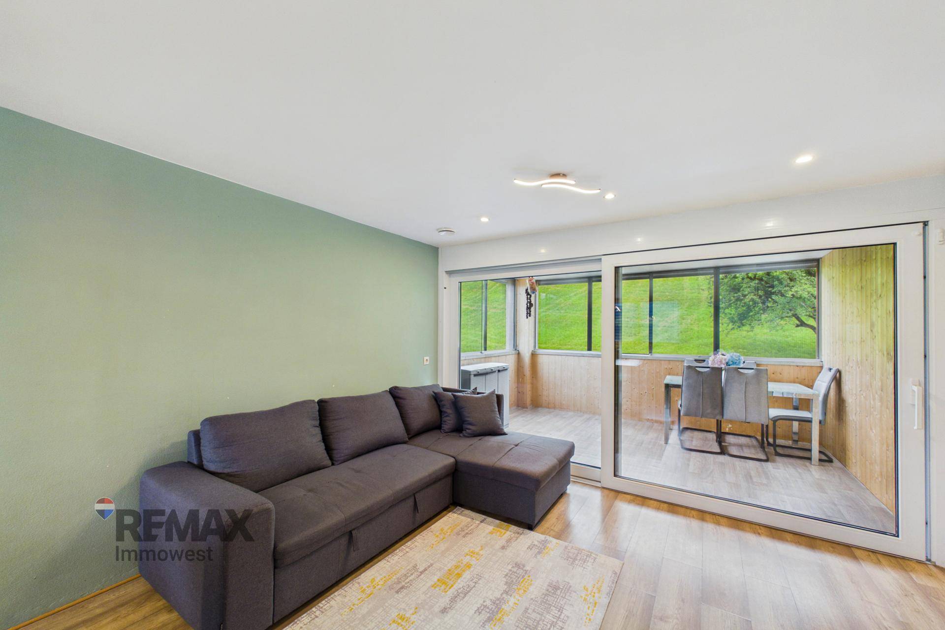 Gemütliches Wohnzimmer mit Ecksofa und Zugang zu einem verglasten Balkon mit grüner Aussicht.