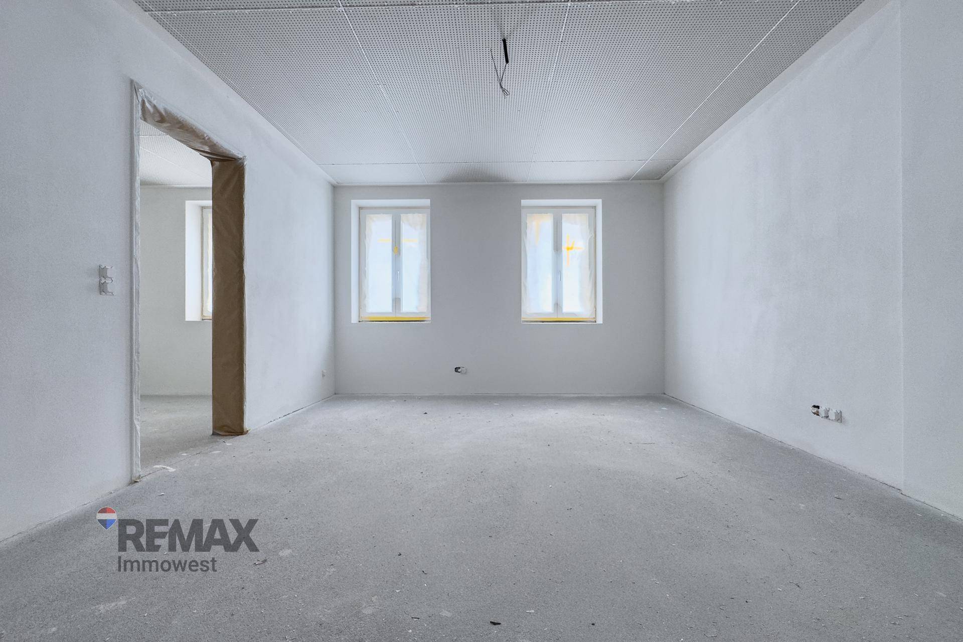 Leerer Raum im Rohbauzustand mit weißen Wänden, Betonboden und zwei verpackten Fenstern.