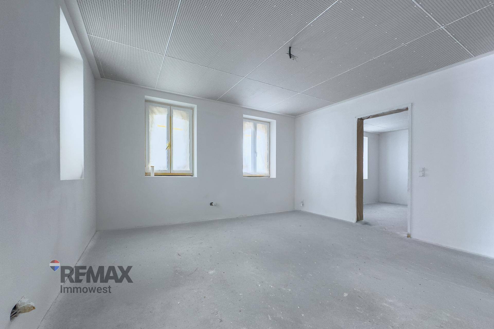 Unmöblierter Raum im Rohbau mit drei Fenstern, Betonboden und abgehängter Decke.