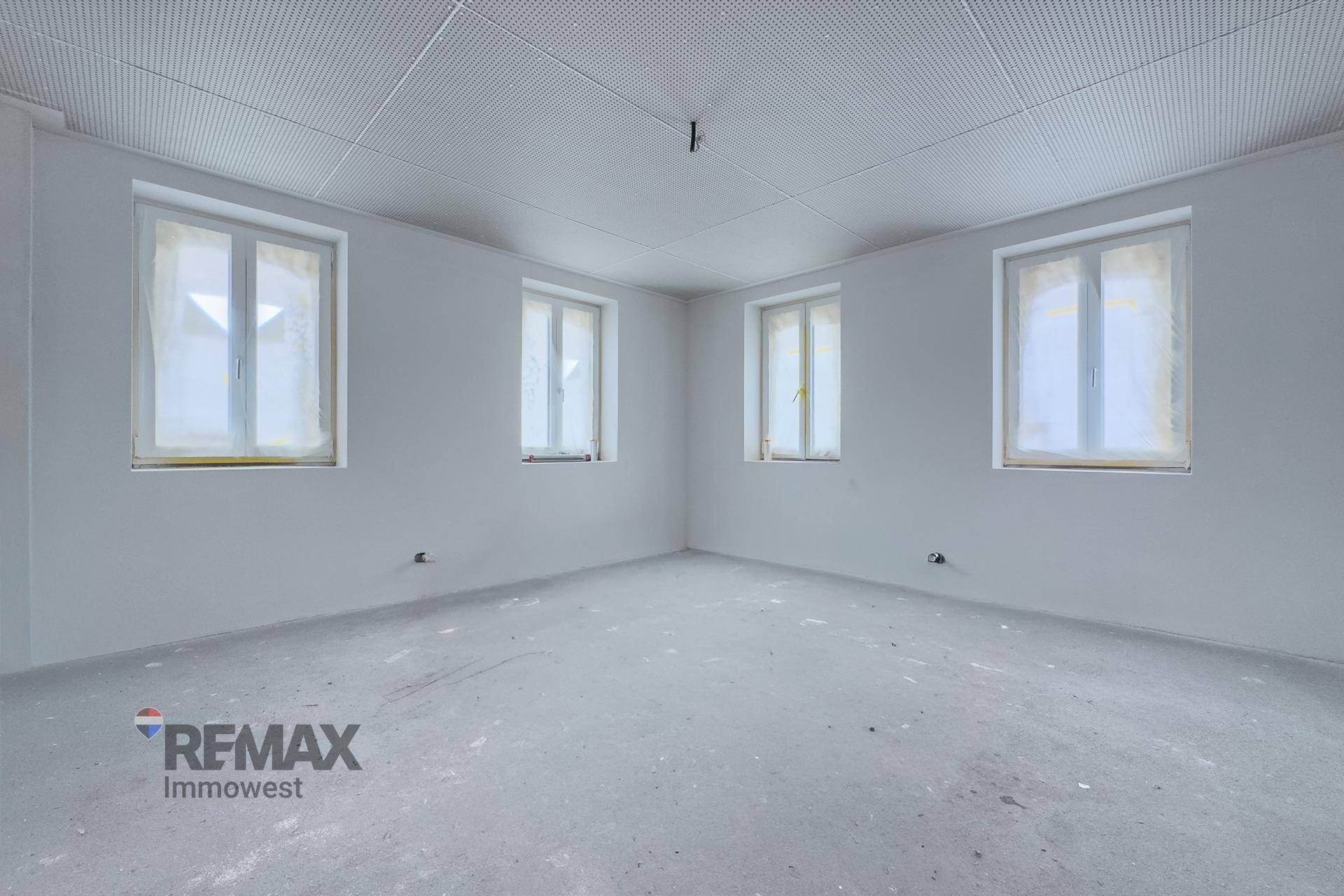 Großer, leerer Raum im Rohbau mit vier Fenstern, Betonboden und Akustikdecke.
