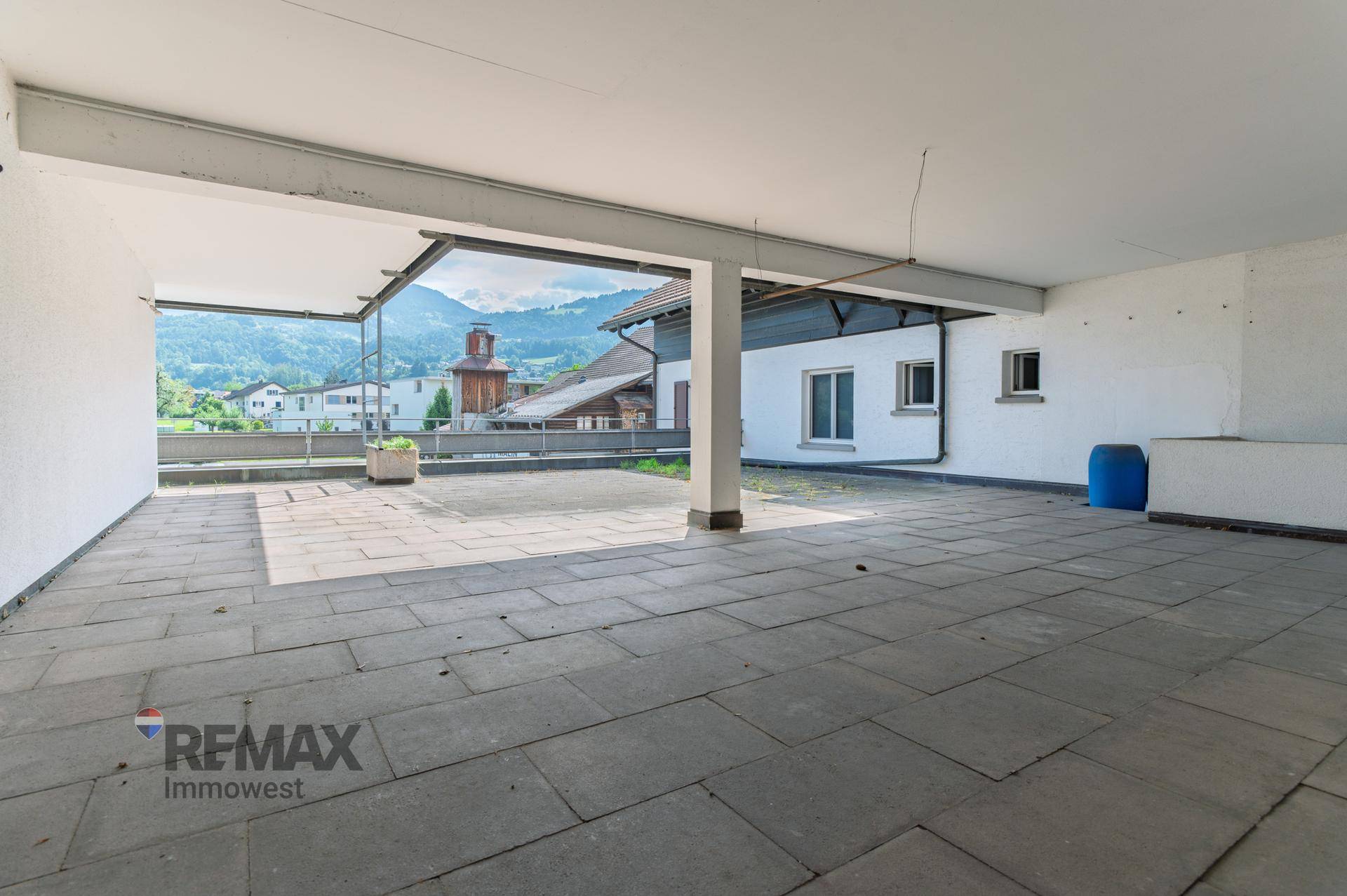 Überdachte Terrasse mit Fliesenboden und Blick auf die Berge und Nachbargebäude.
