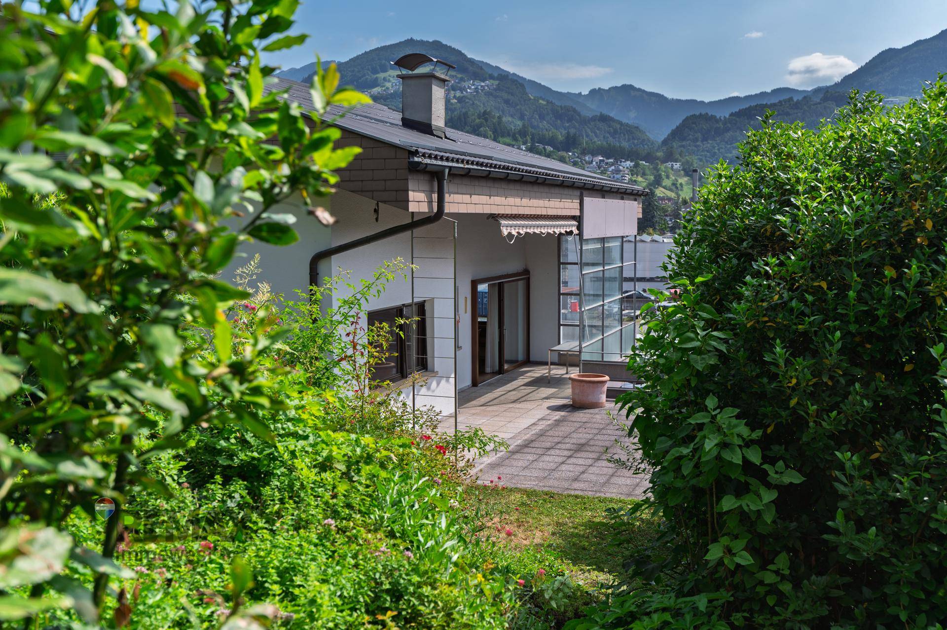 Hausfassade mit Terrasse, umgeben von üppigem Grün und Blick auf die Berge.