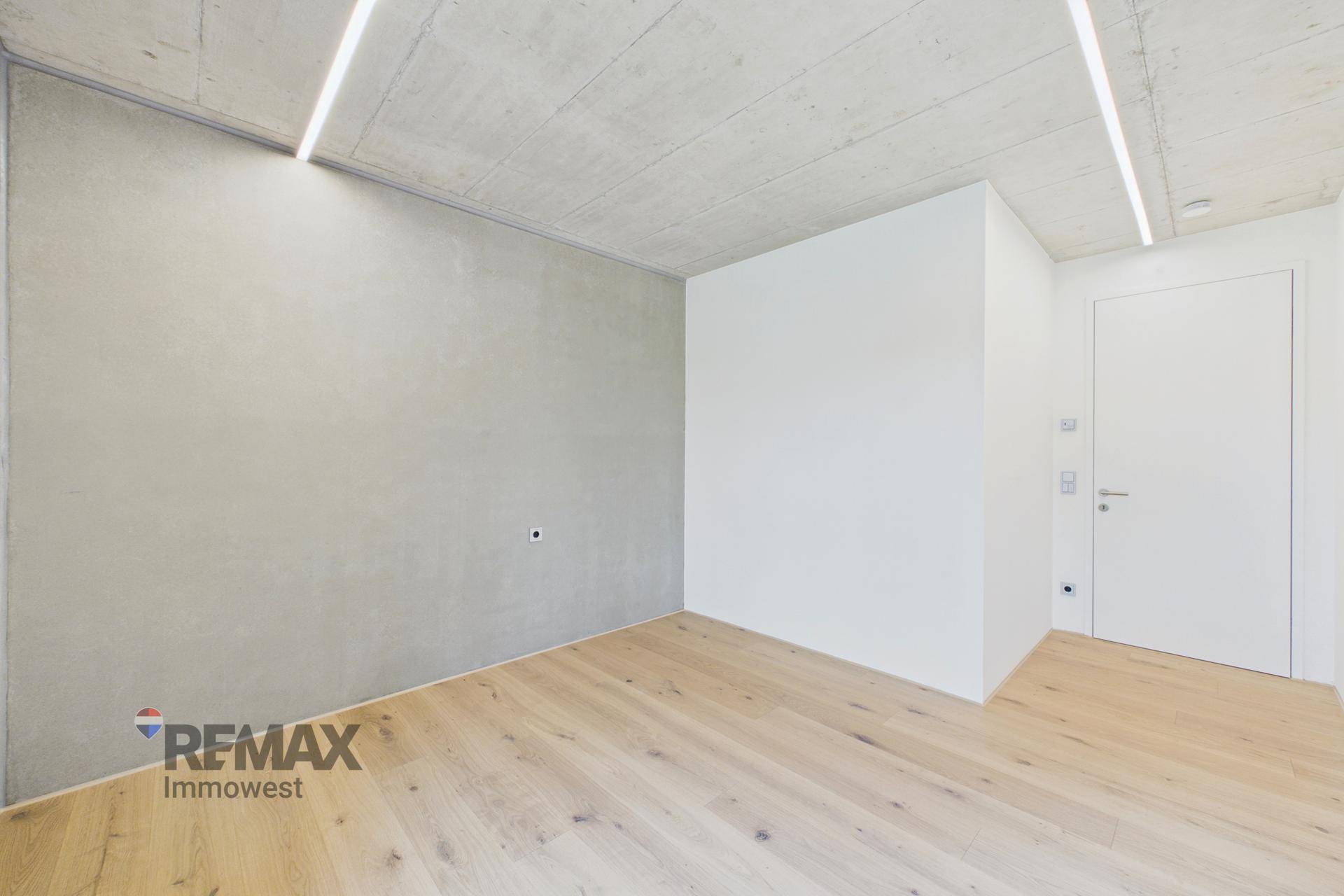 Helles Zimmer mit Holzboden, Sichtbetondecke und weiß gestrichenen Wänden.